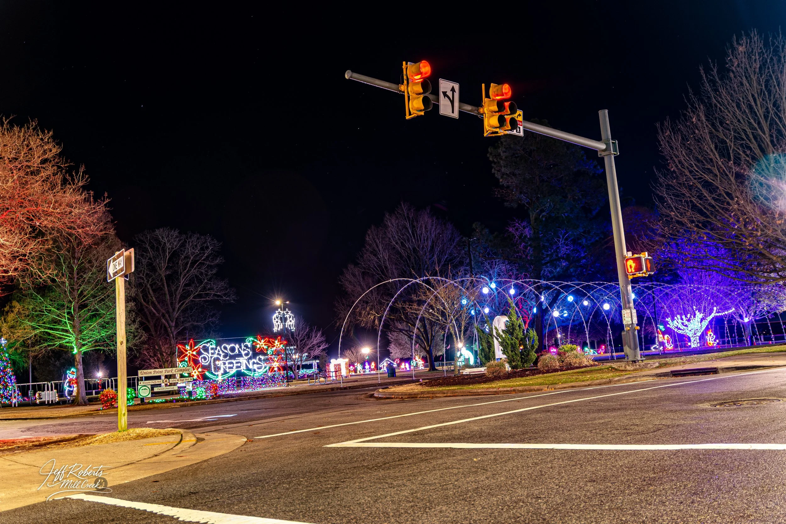 Night view of a park decorated with colorful Christmas lights and signs that say 'Season's Greetings,' with empty streets and traffic signals.