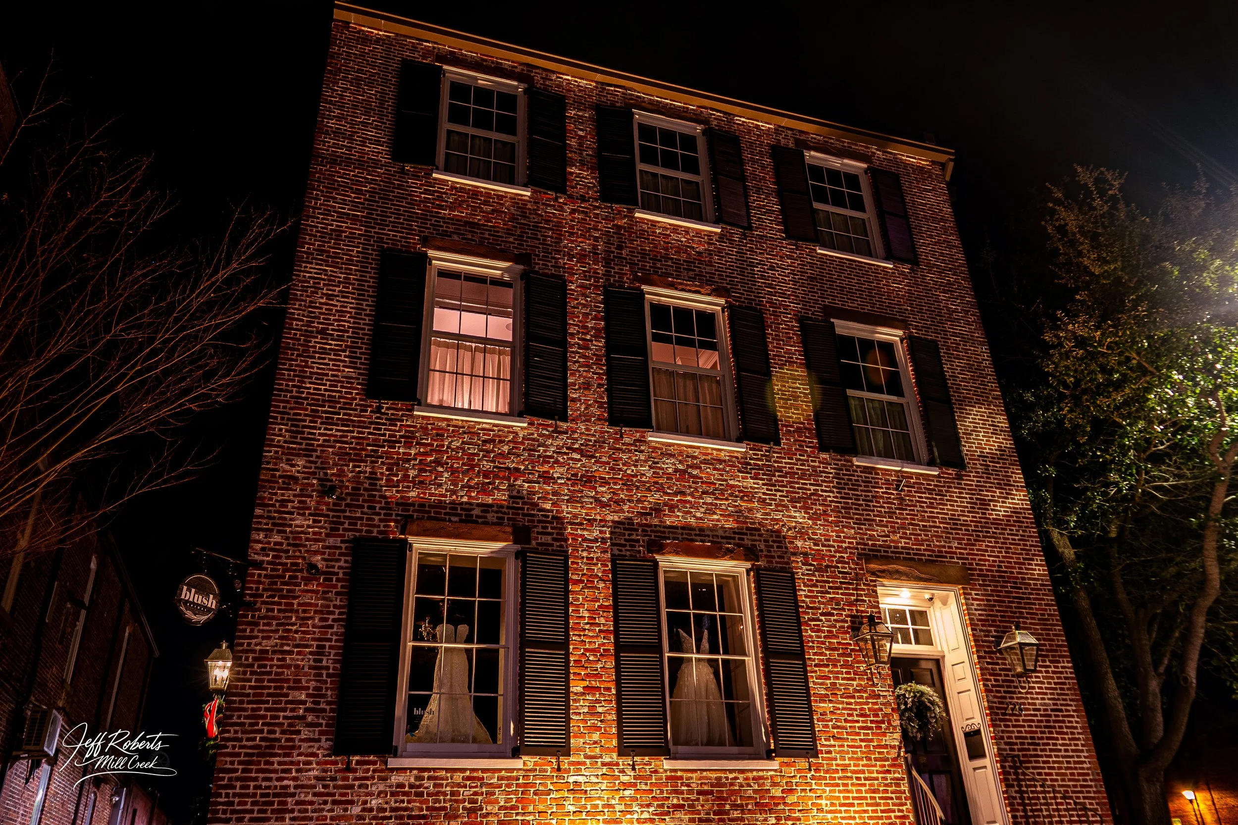 A brick four-story building at night with lit windows, black shutters, and a decorated entrance door.