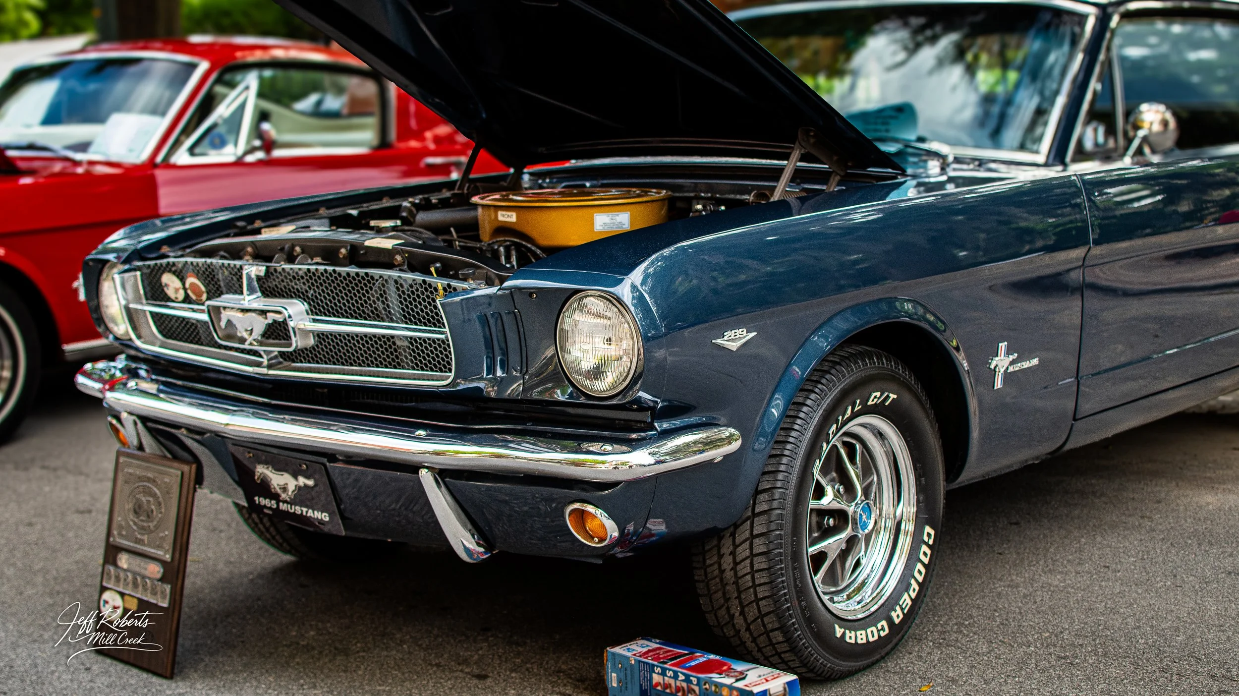 A vintage dark blue 1965 Ford Mustang fastback car with its hood open, showing the engine. Other classic cars are visible in the background at an outdoor car show.