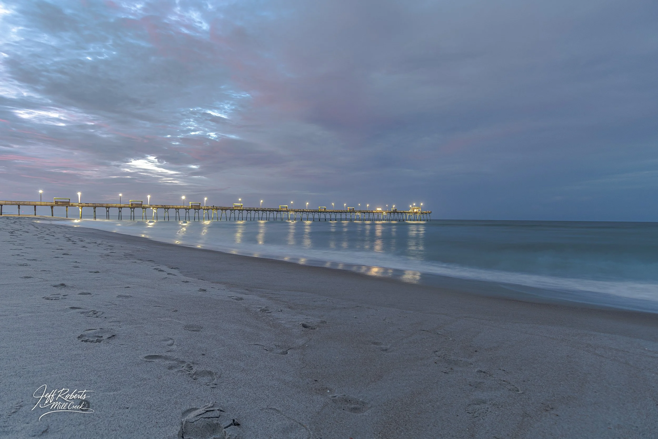 Long pier extending into the ocean at dusk with reflection of lights on water, sandy beach in foreground, cloudy sky above.