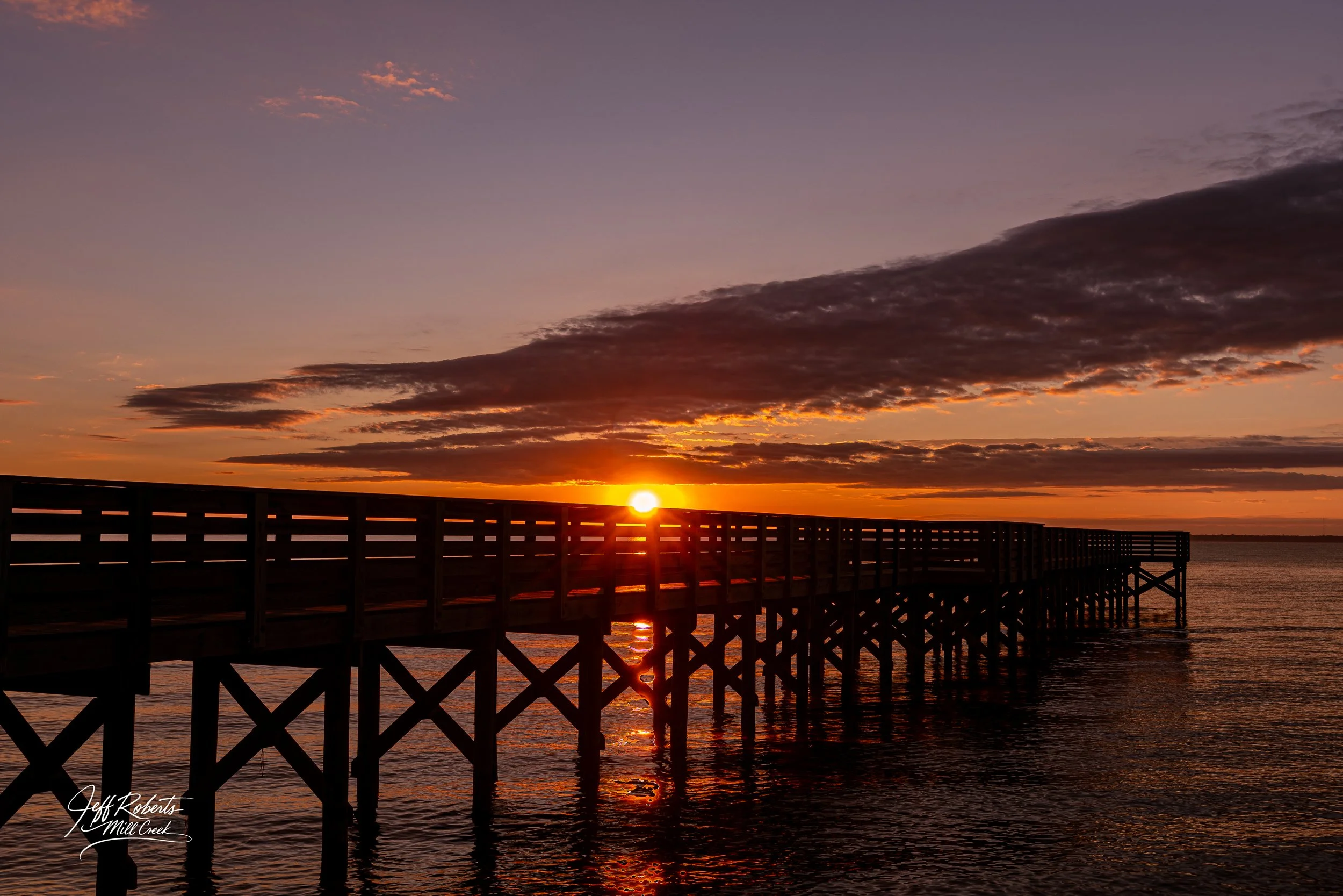 Sunset over a wooden pier extending into the water with clouds in the sky.