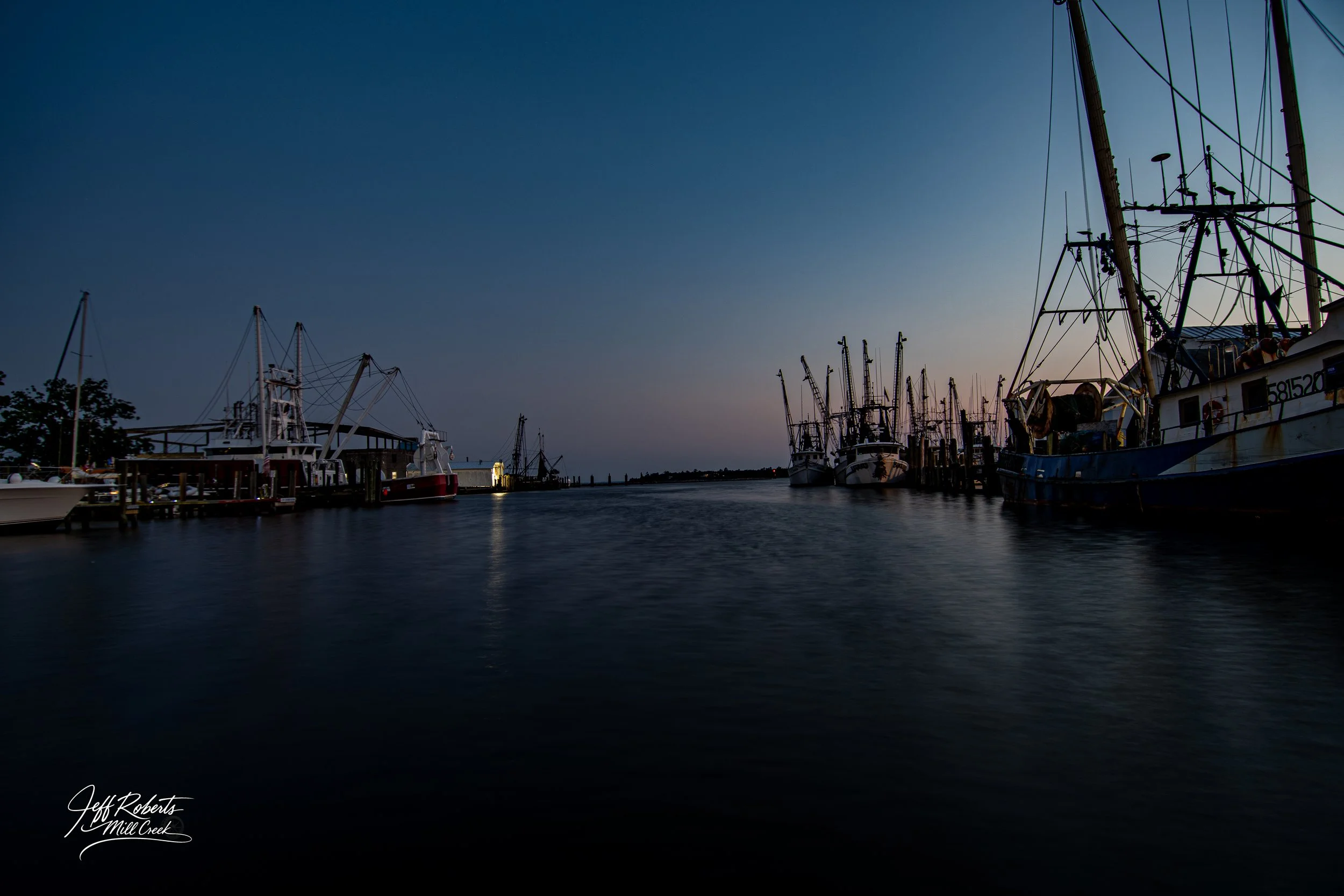 Marina with several docked fishing boats during dusk, with the sky transitioning from blue to purple, and calm water reflecting the boats.