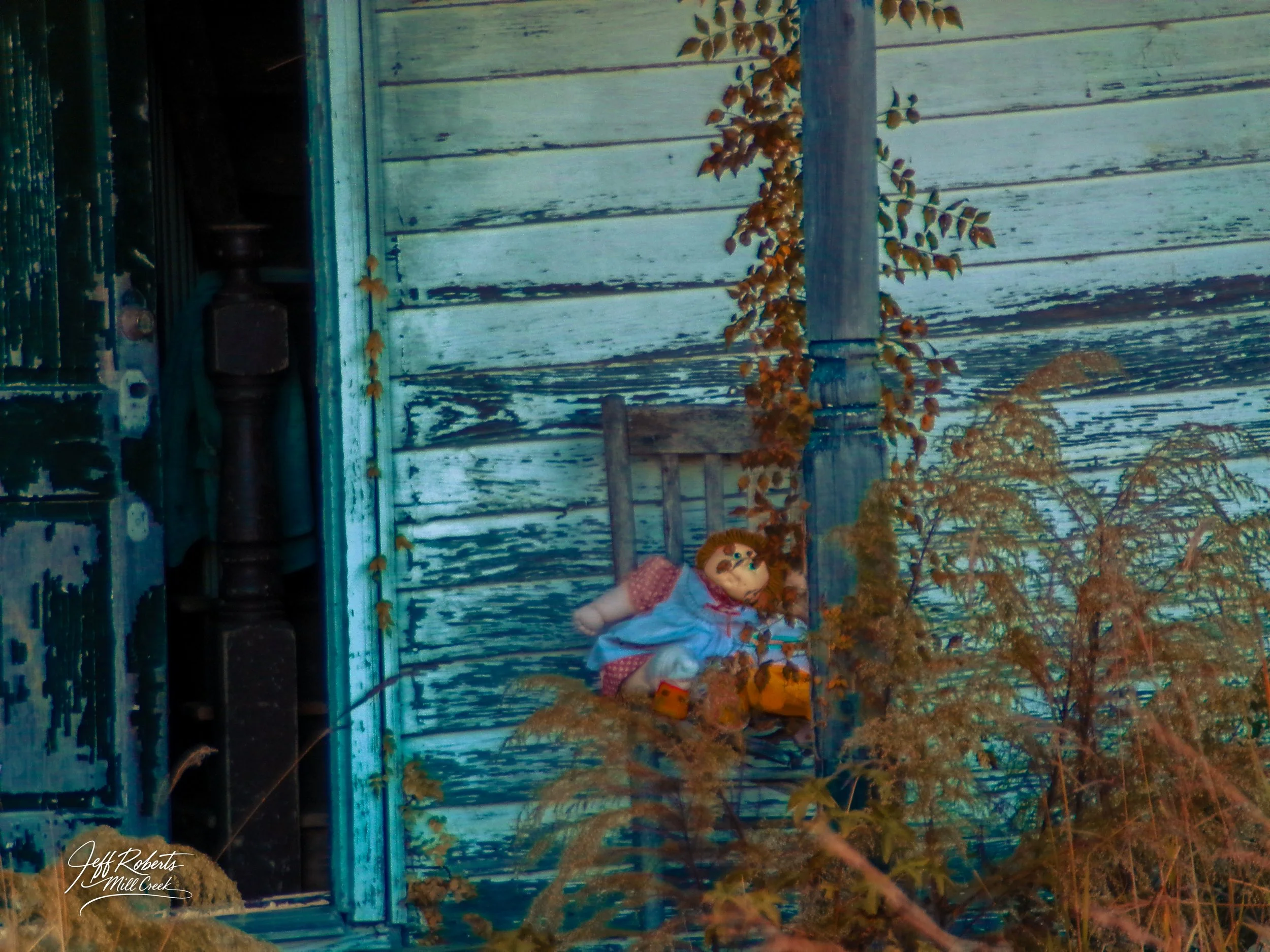 A doll with brown hair, red cheeks, wearing a blue jacket, red-patterned dress, and yellow shoes, sitting on a porch next to a weathered blue wooden wall with dried plants around.