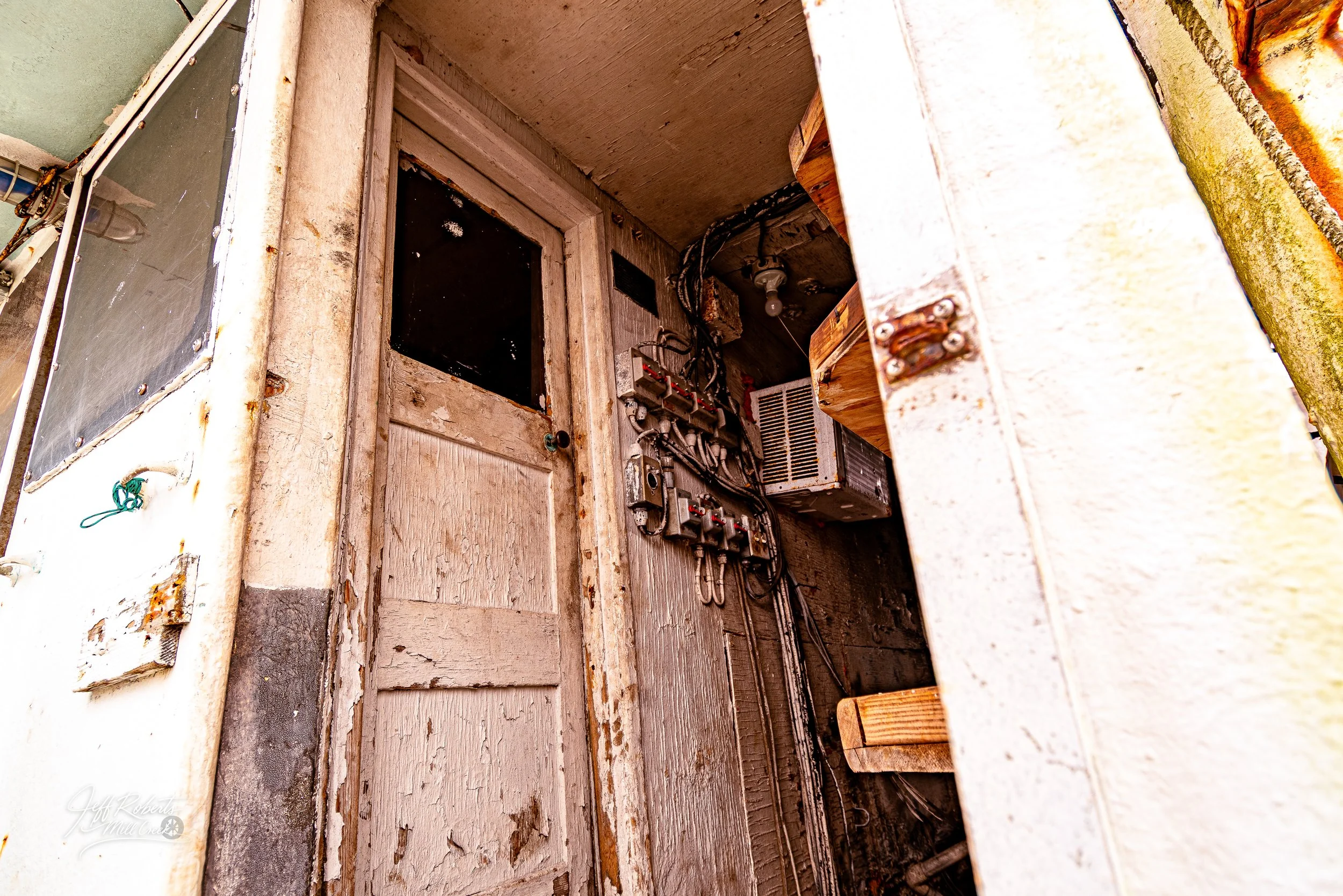 Old, weathered door with chipped paint and electrical wiring on the wall, next to a small window and a wooden shelf, in a rustic or abandoned space.