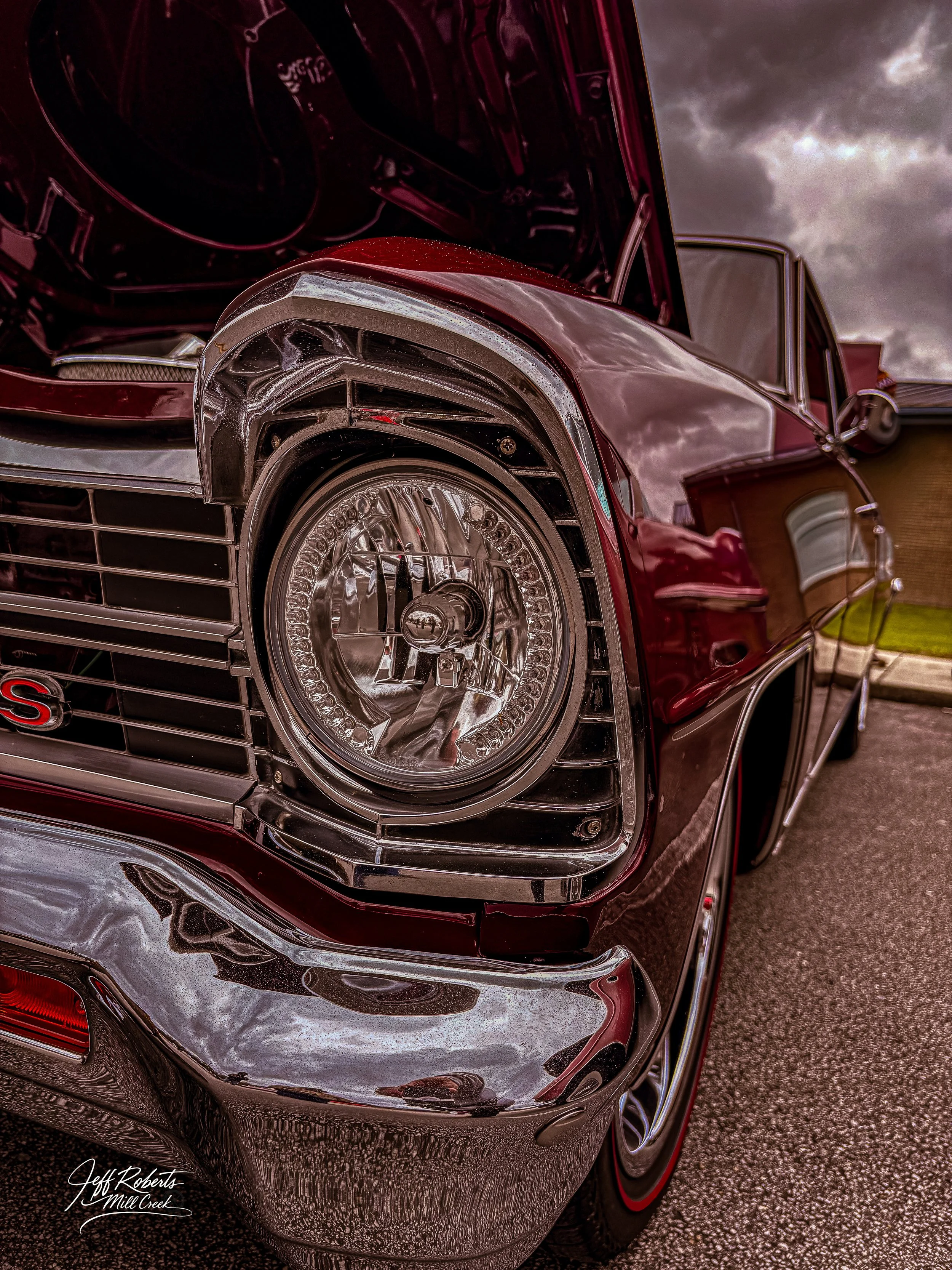 Close-up of a vintage red car's front left side, showing a round headlight, chrome bumper, and part of the open hood with engine components inside. The sky is cloudy in the background.