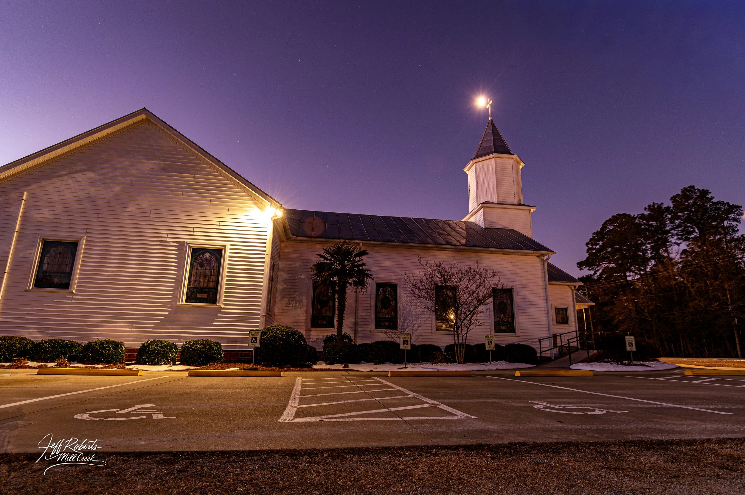 Church building at night with parking lot in foreground, handicap parking spaces, small trees, and a clear starry sky with a few visible stars.