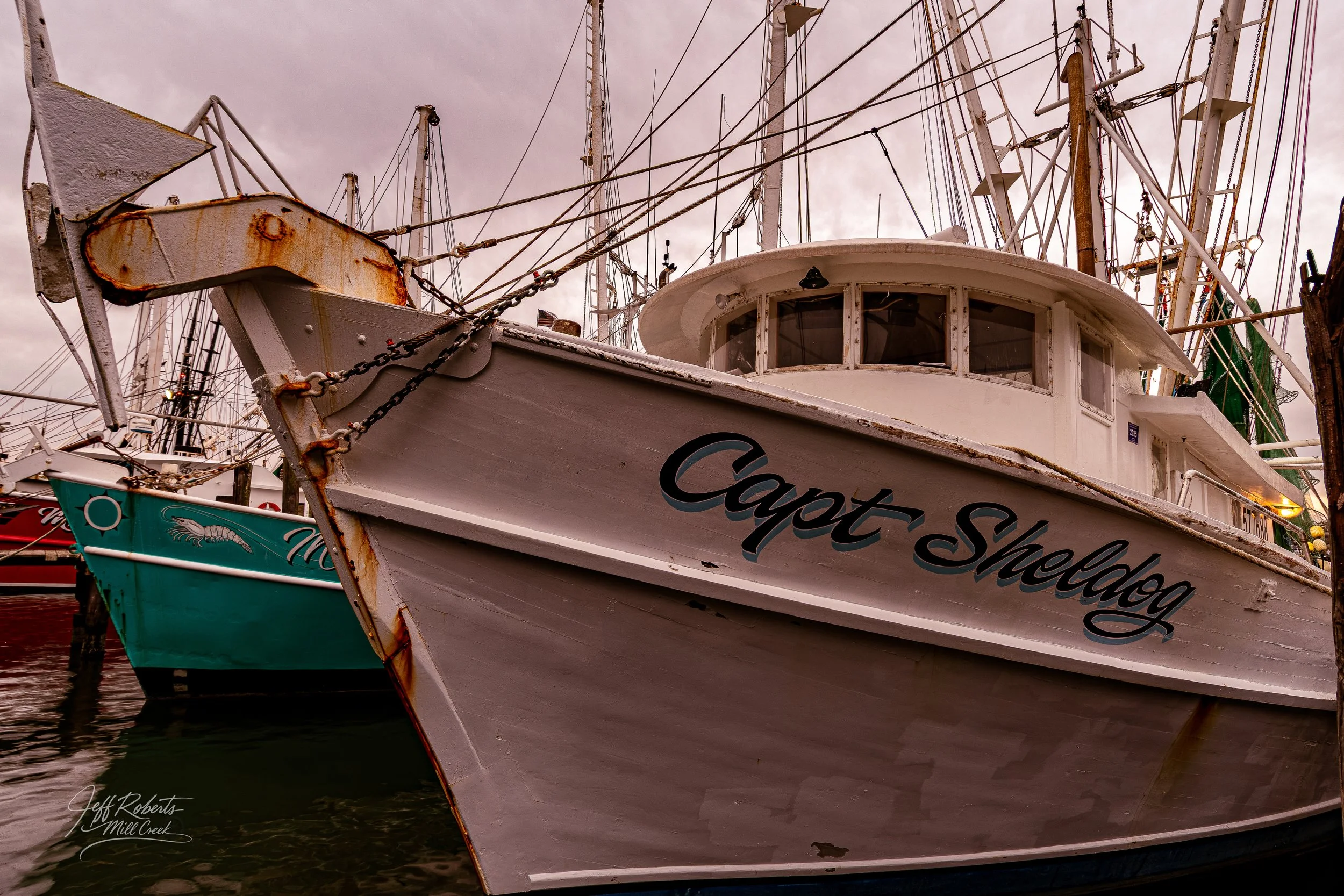 Close-up of a white boat named "Capot Shelly" docked at a marina, with other boats and masts in the background under cloudy sky.