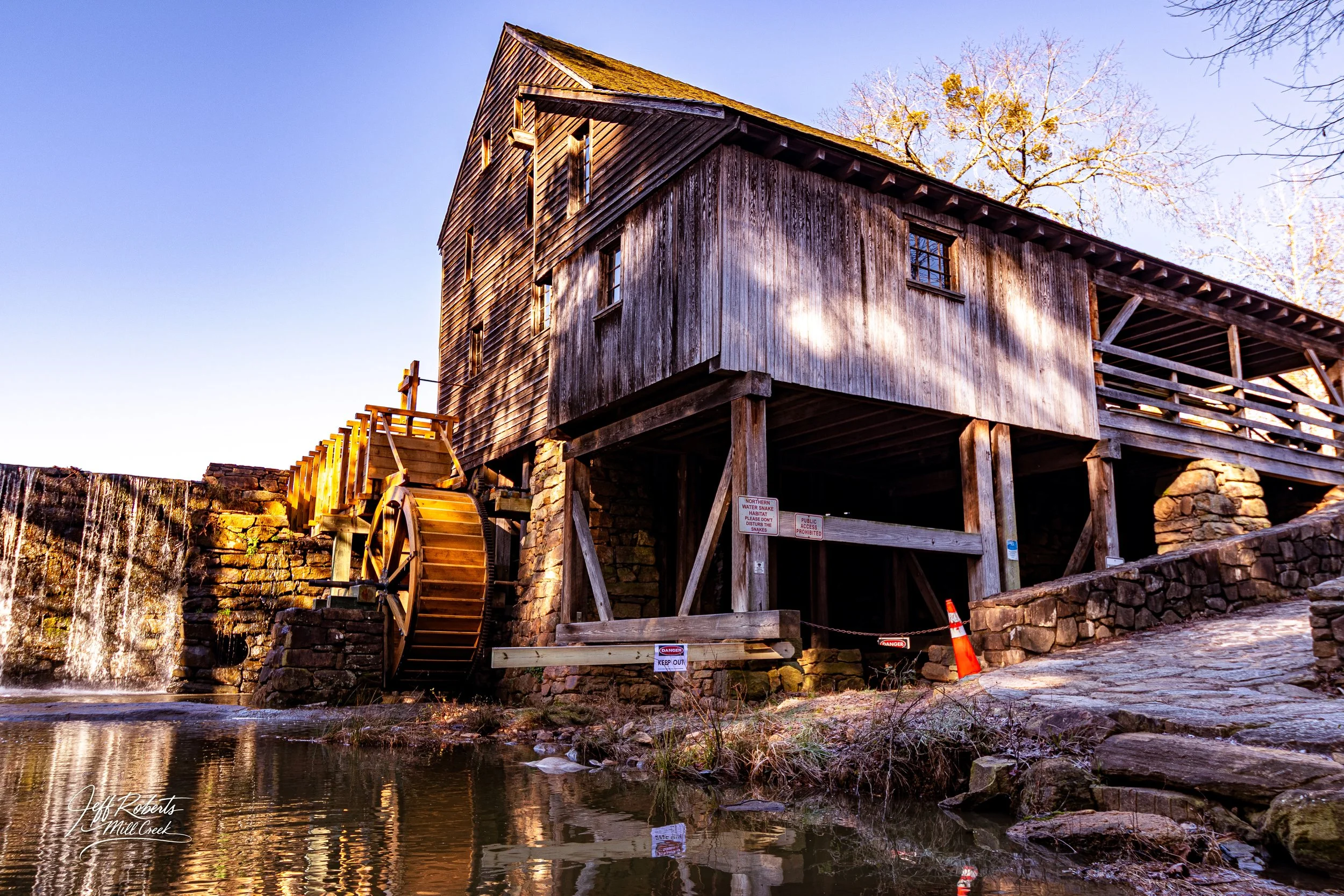An old wooden mill with a waterwheel, situated next to a small waterfall and water, with signs and a traffic cone nearby.