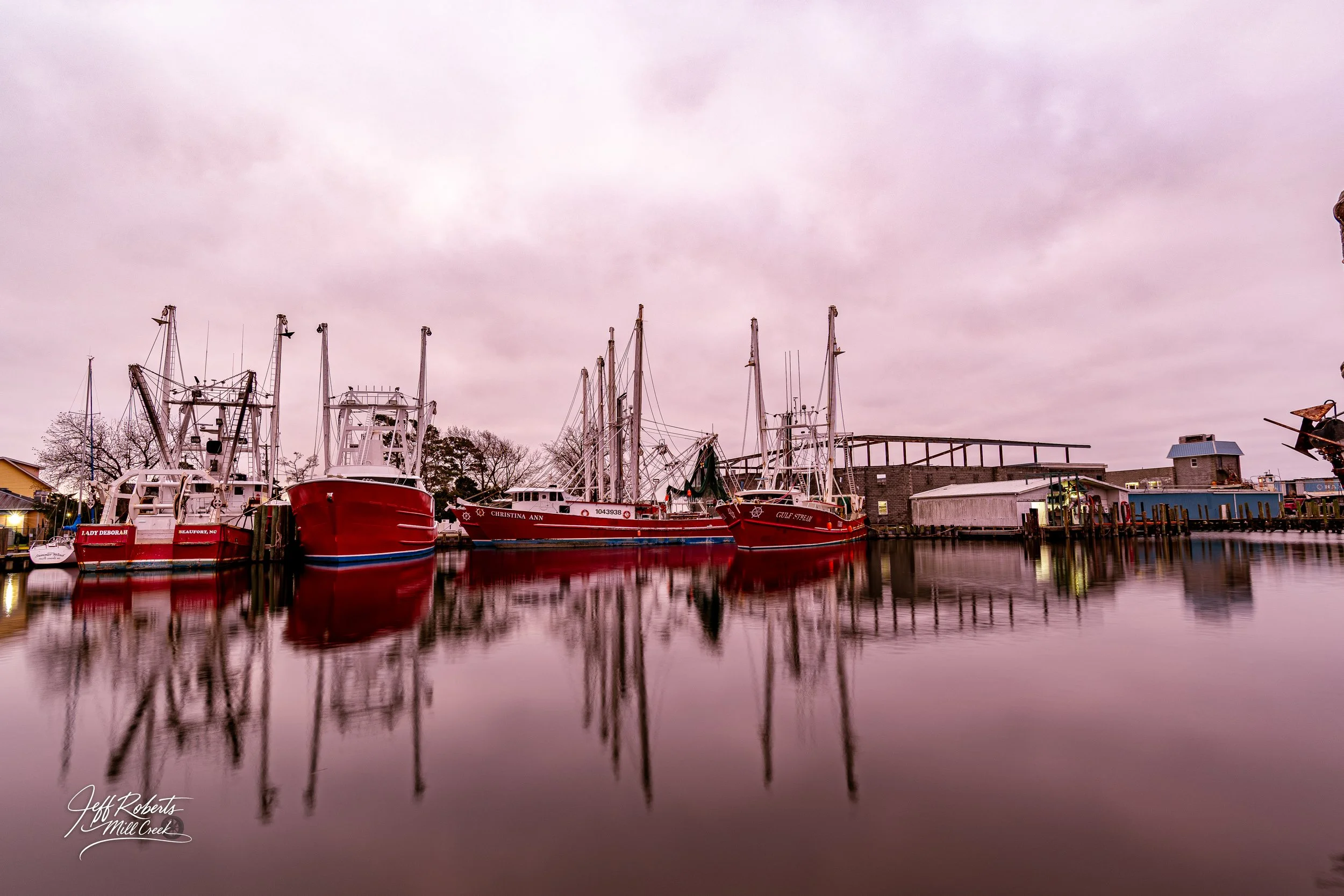 Boats docked at a marina with reflections on calm water, cloudy sky, and buildings in the background.