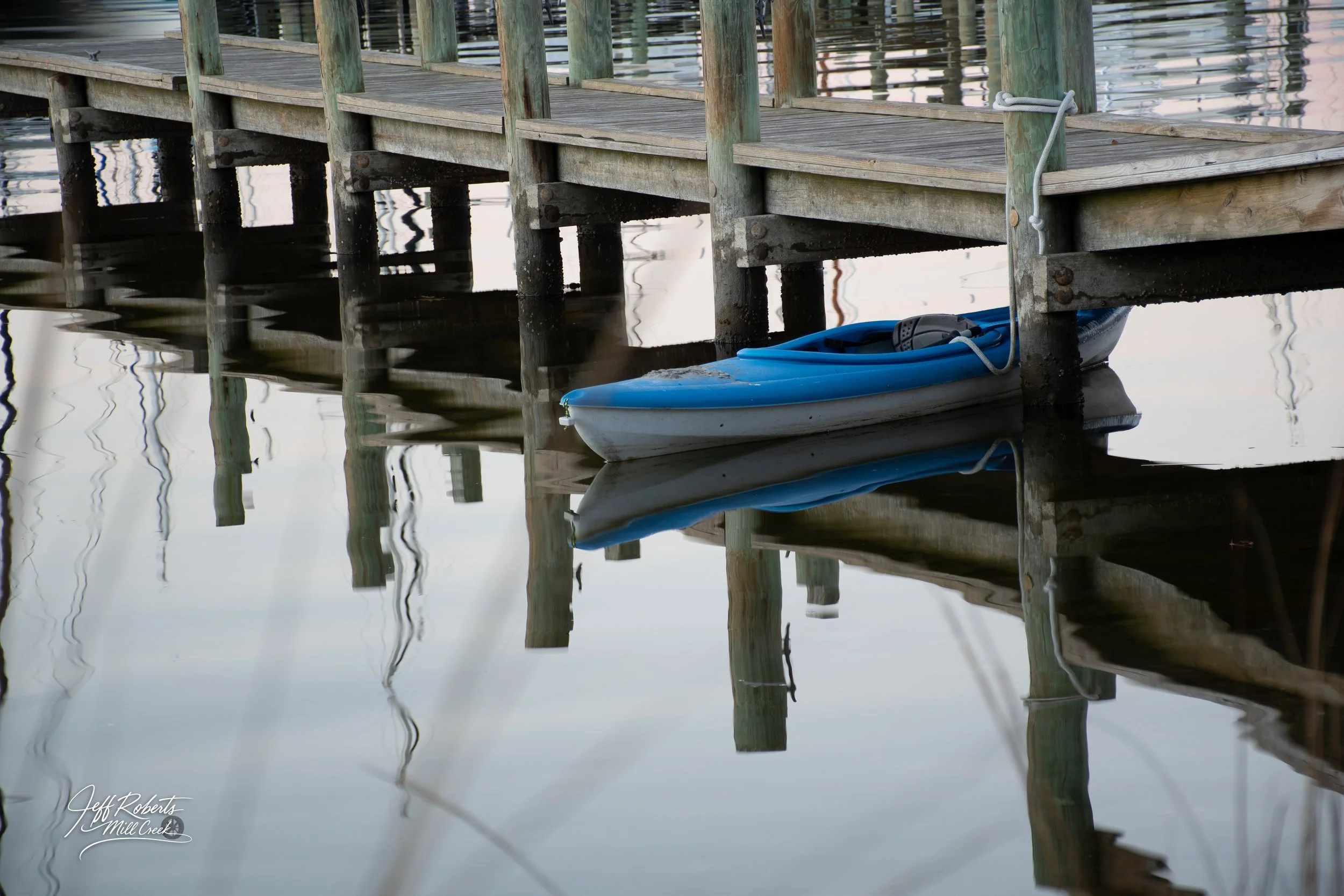 A wooden dock extending over calm water, with a blue kayak tied to the dock and reflecting in the water.
