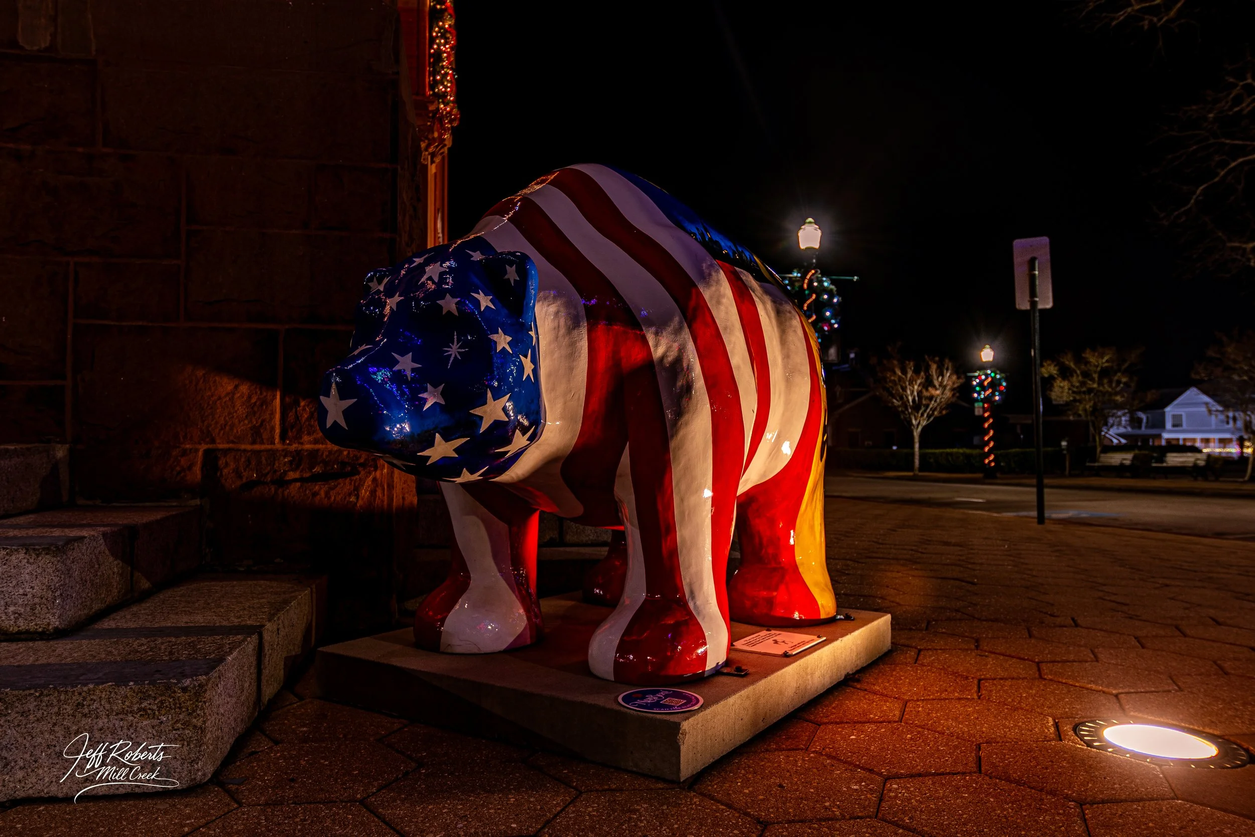 A polar bear statue painted with the American flag design, situated outdoors at night, with Christmas lights and street lamps in the background.