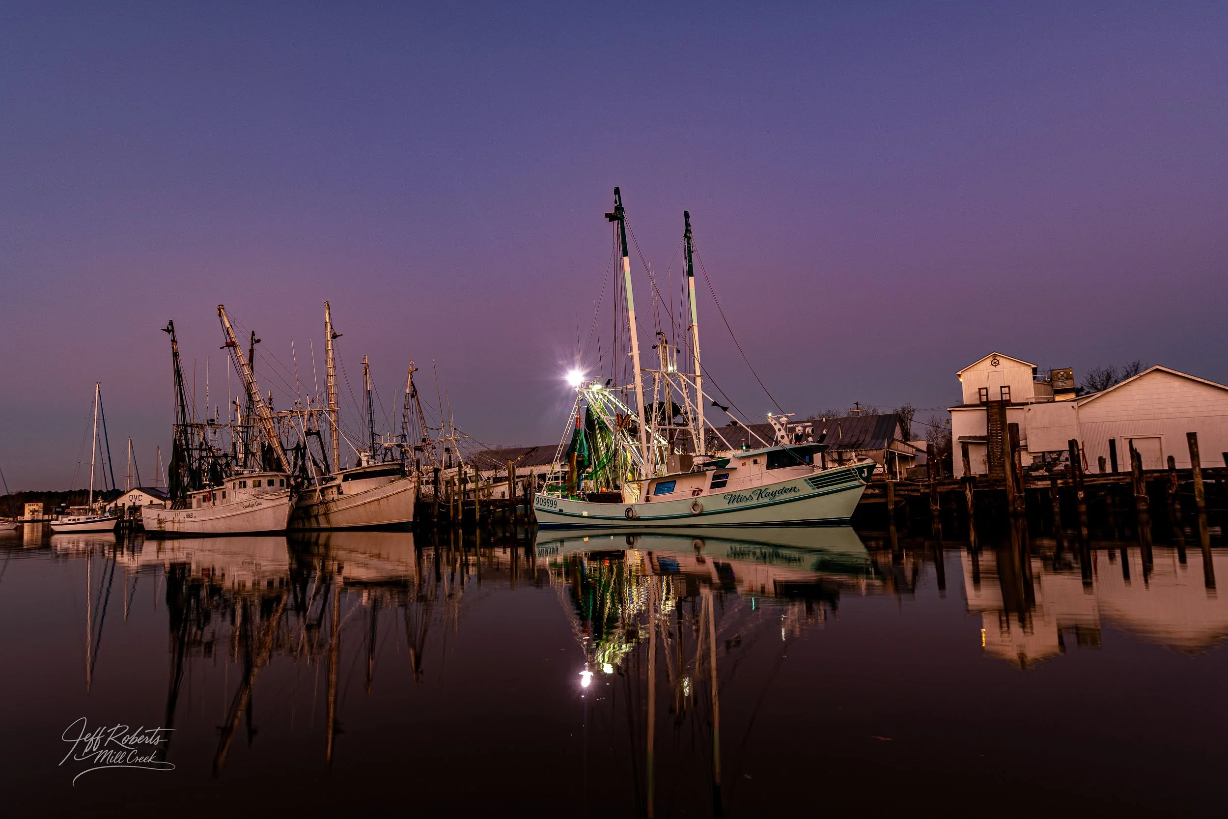 A group of boats docked at a marina during twilight with reflections in the water and a purple sky.