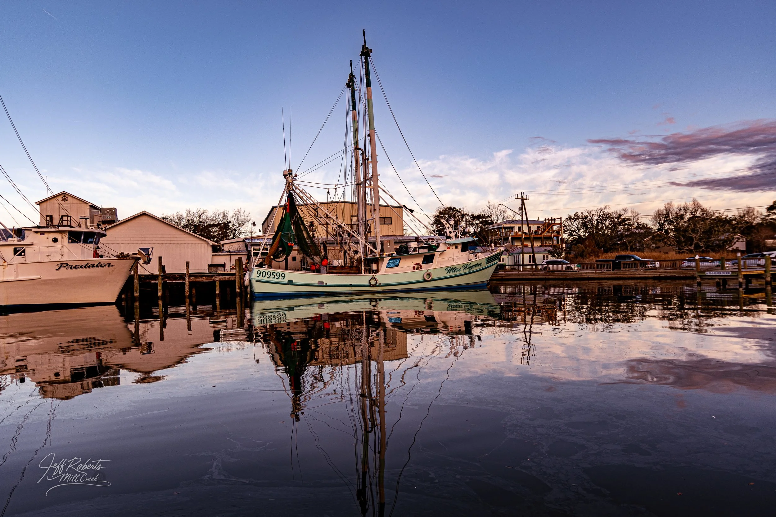 A boat docked at a marina during sunset with reflections in the water, houses, and trees in the background.