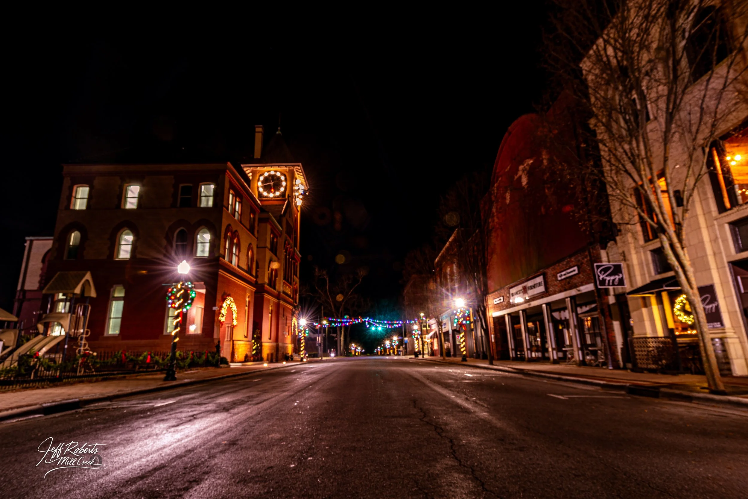 Empty street at night decorated with Christmas lights, wreaths, and candy cane street lamps, with a historic building on the left and modern shops on the right.