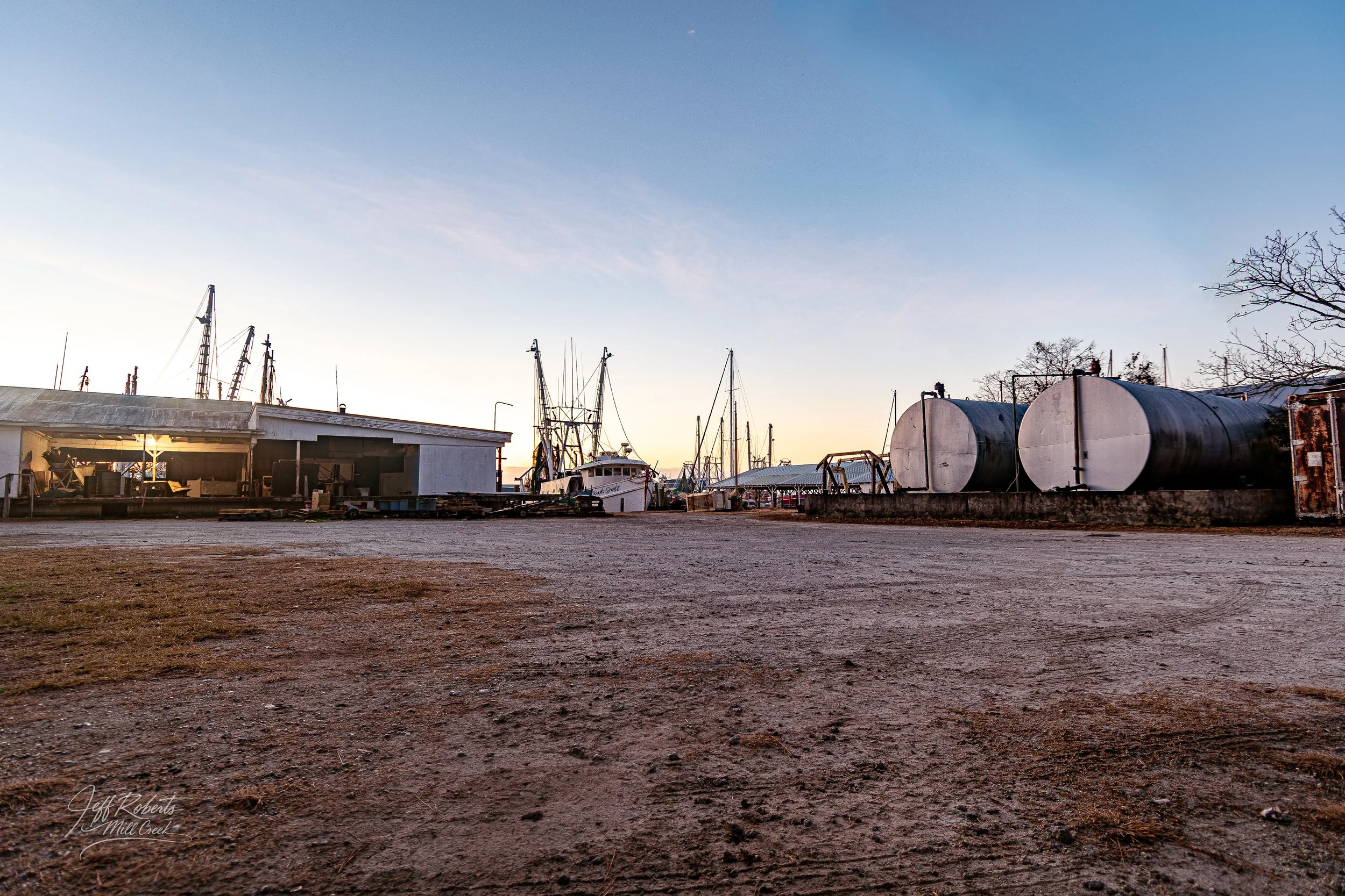 A boatyard with boats, storage tanks, and a building at sunset, with a dirt ground in the foreground.