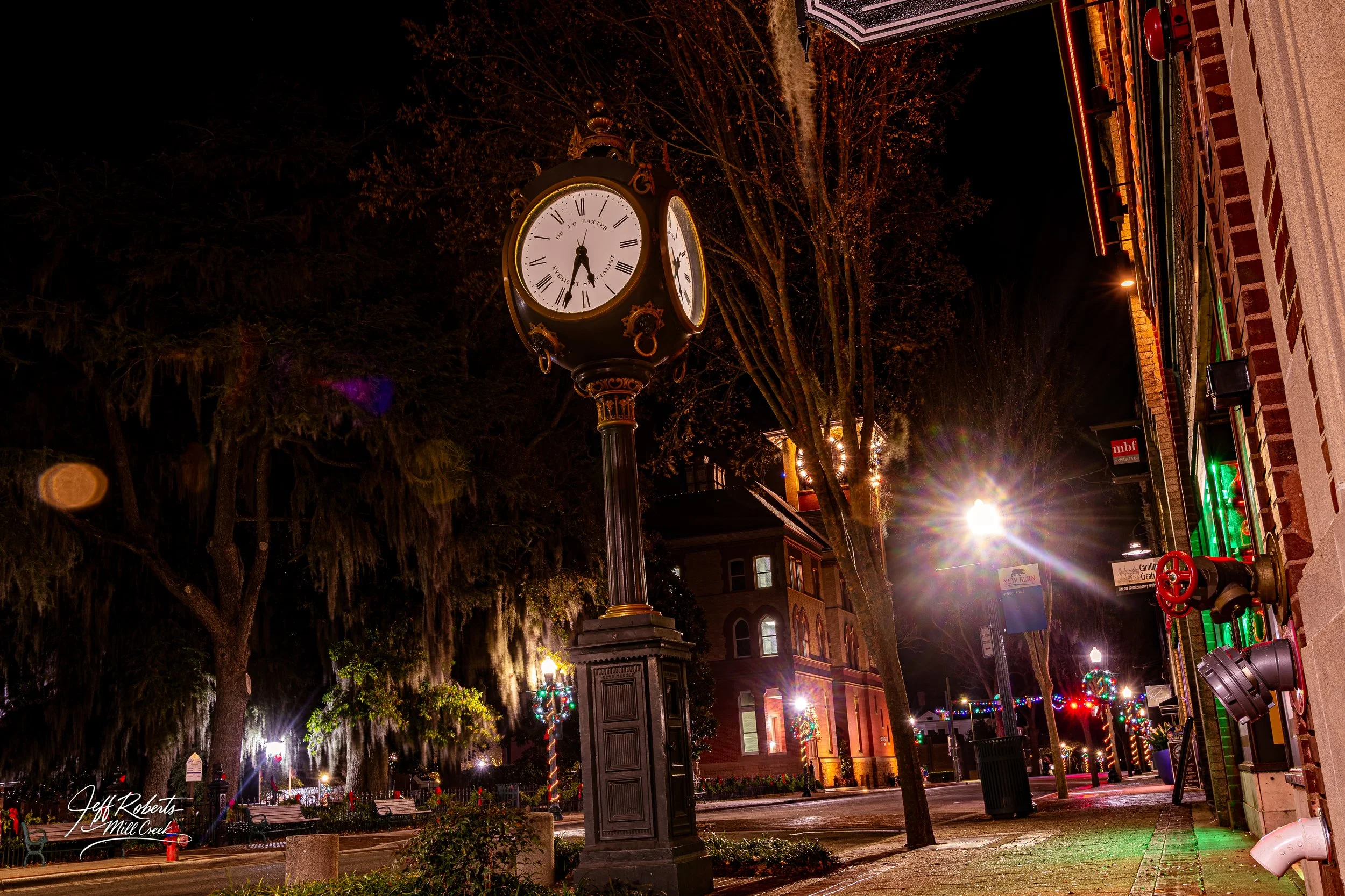 Nighttime street scene with an ornate clock on a tall black lamppost, decorated with gold accents, showing the time at 9:23. Trees draped with Spanish moss line the sidewalk, and buildings with festive holiday lights are visible. Bright streetlights 