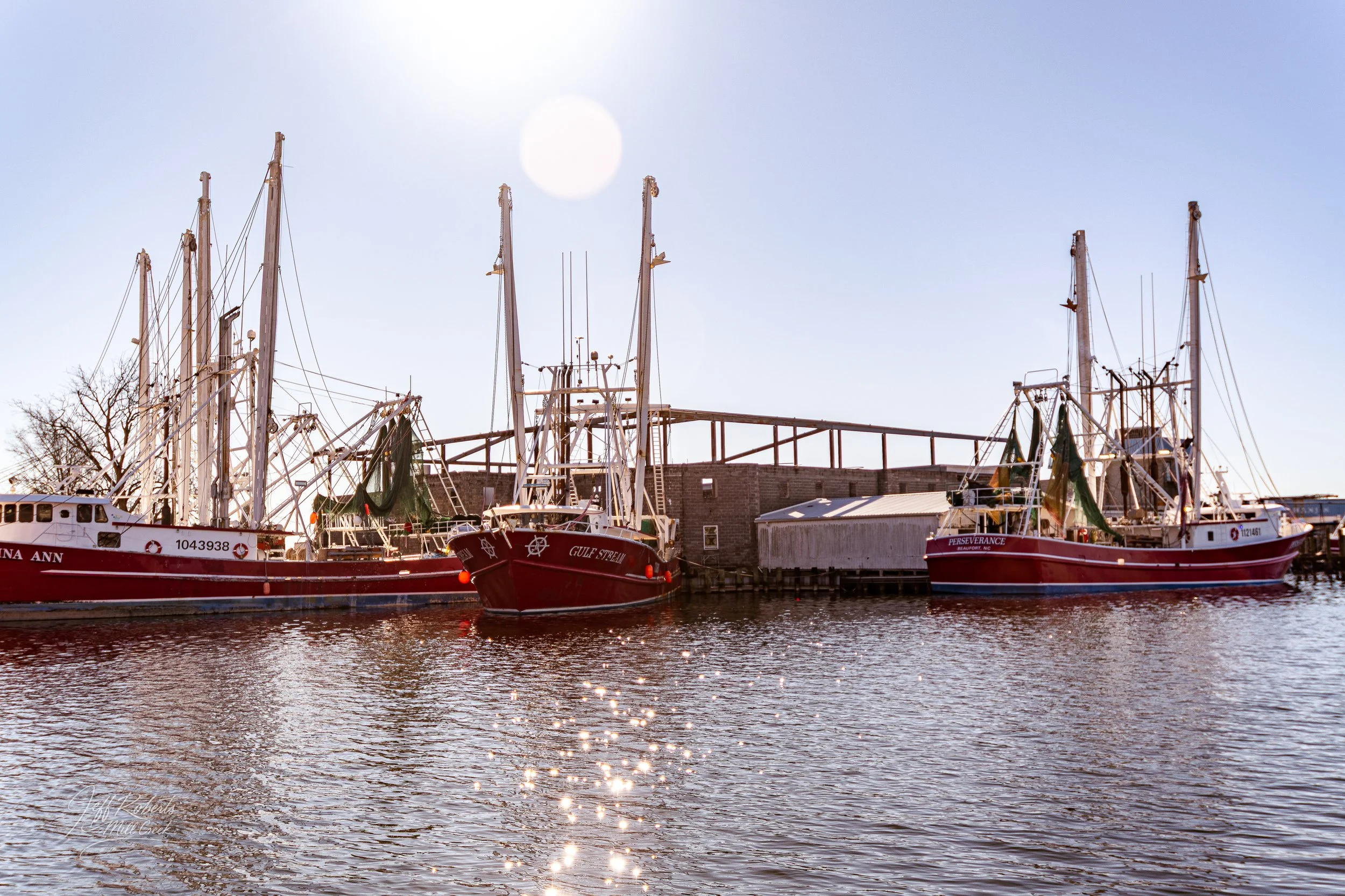 Three red and white fishing boats docked at a pier on calm water with small ripples, under a clear sky with bright sunlight reflecting on the water.