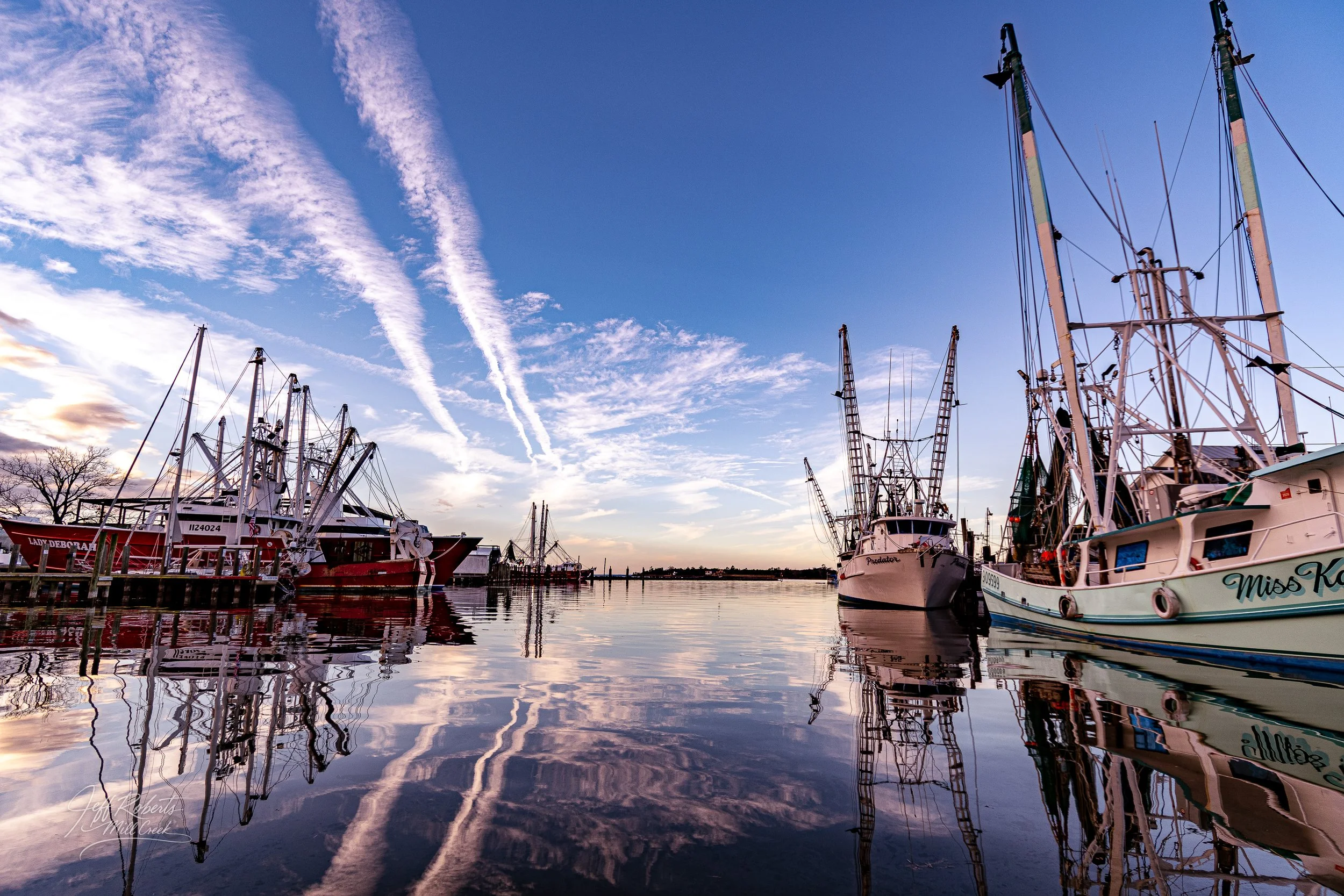 A serene harbor scene with several docked fishing boats reflecting in calm water during sunset, with a blue sky and wispy clouds overhead.