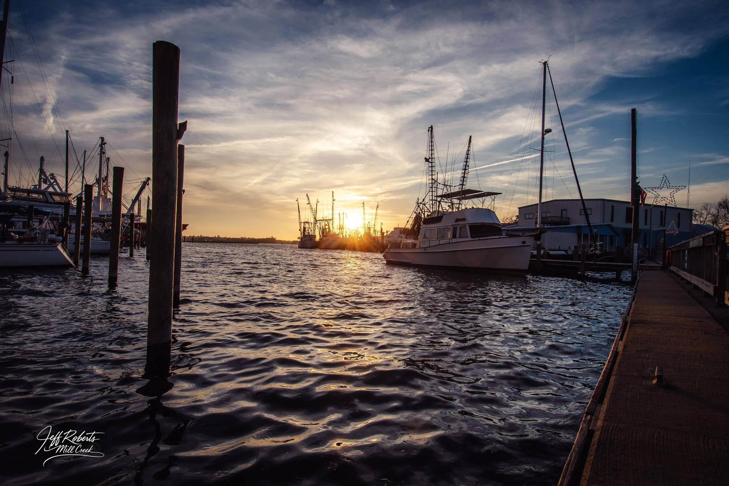 Boats docked at a marina during sunset with a bridge and colorful sky in the background.
