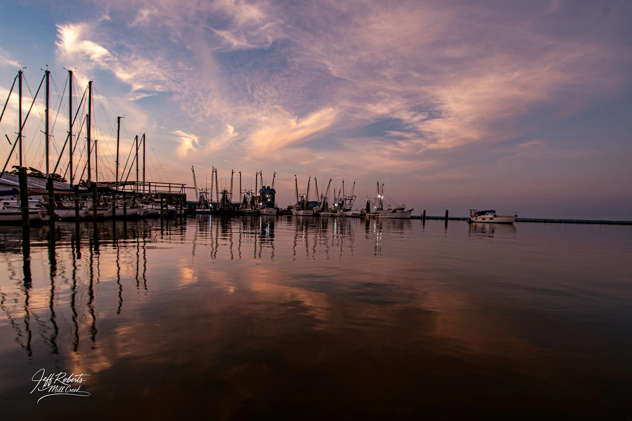 A harbor at sunset with sailboats docked, calm water reflecting the sky and boats, pink and purple clouds.
