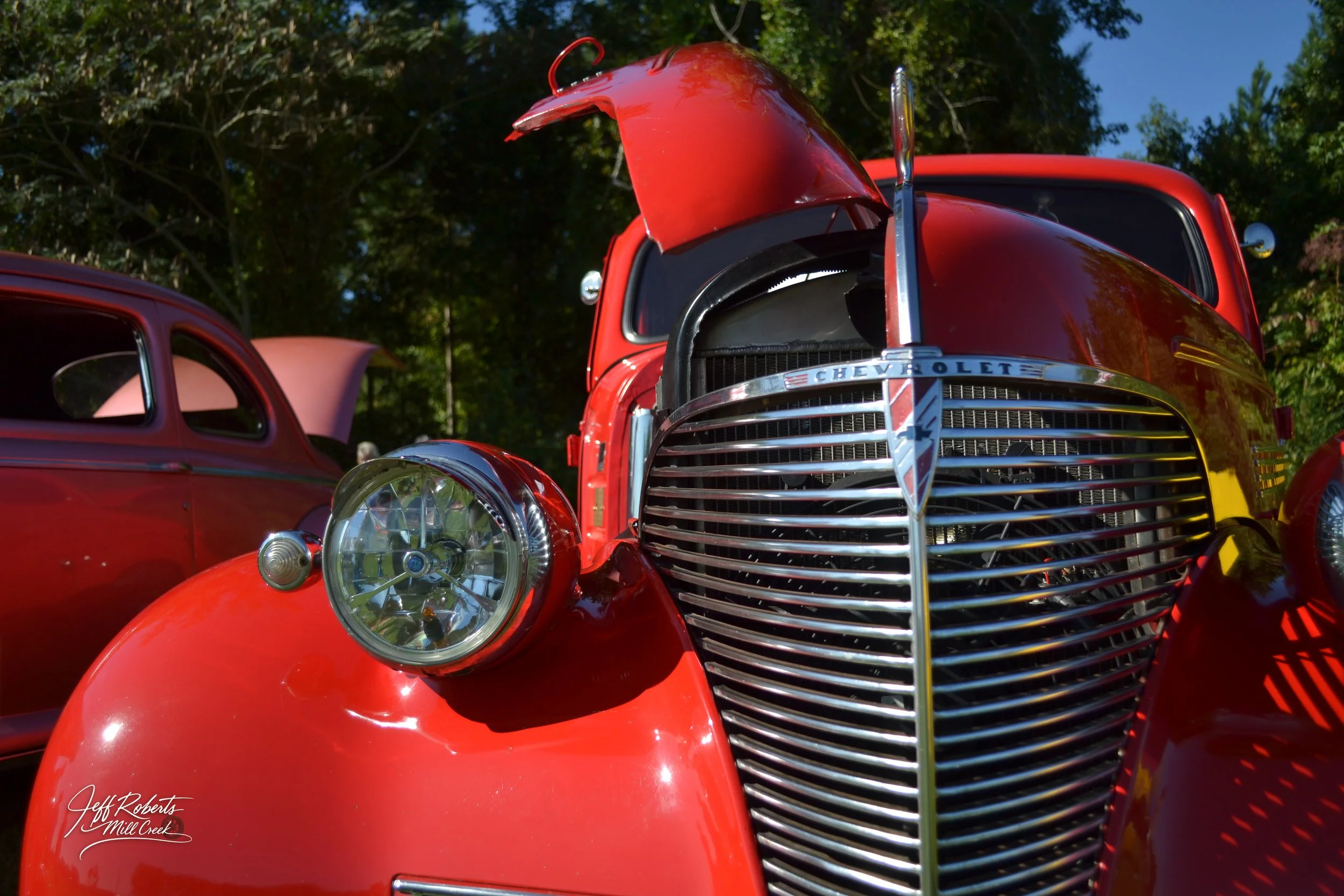 Close-up of front of vintage red Chevrolet car with hood open, showing the grille and headlight, parked outdoors with trees in background.