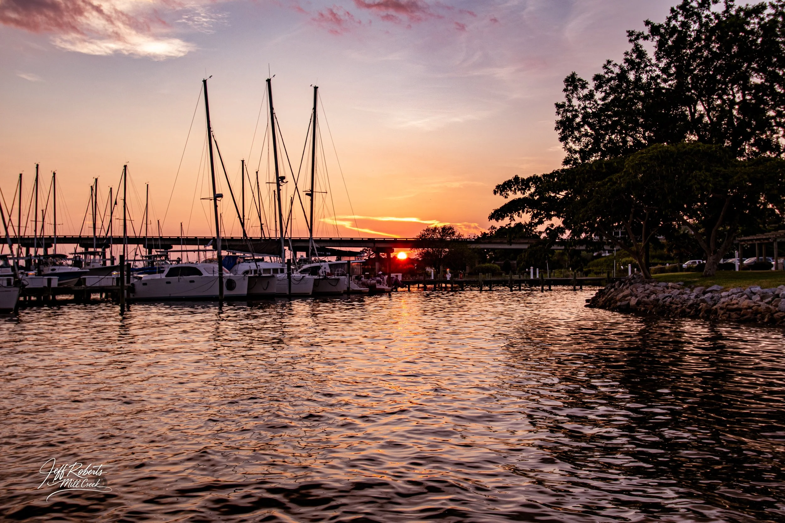 Sailboats docked at a marina during sunset with a sky in shades of pink and orange, trees on the right and water reflecting the sunset colors.