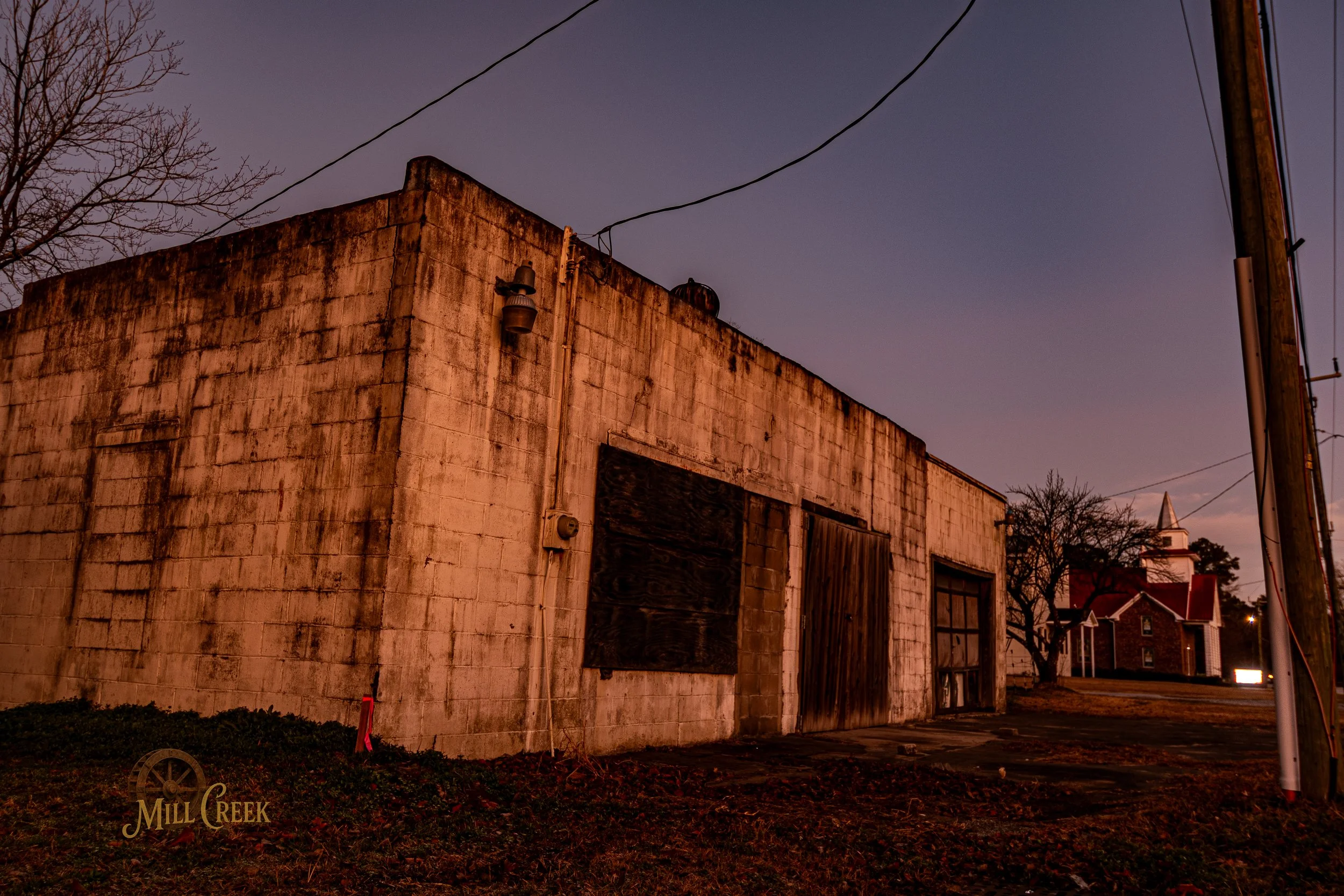 An old, weathered brick building with boarded-up windows and a wooden door, under a twilight sky with leafless trees in the background. The Mill Creek logo is visible in the lower left corner.