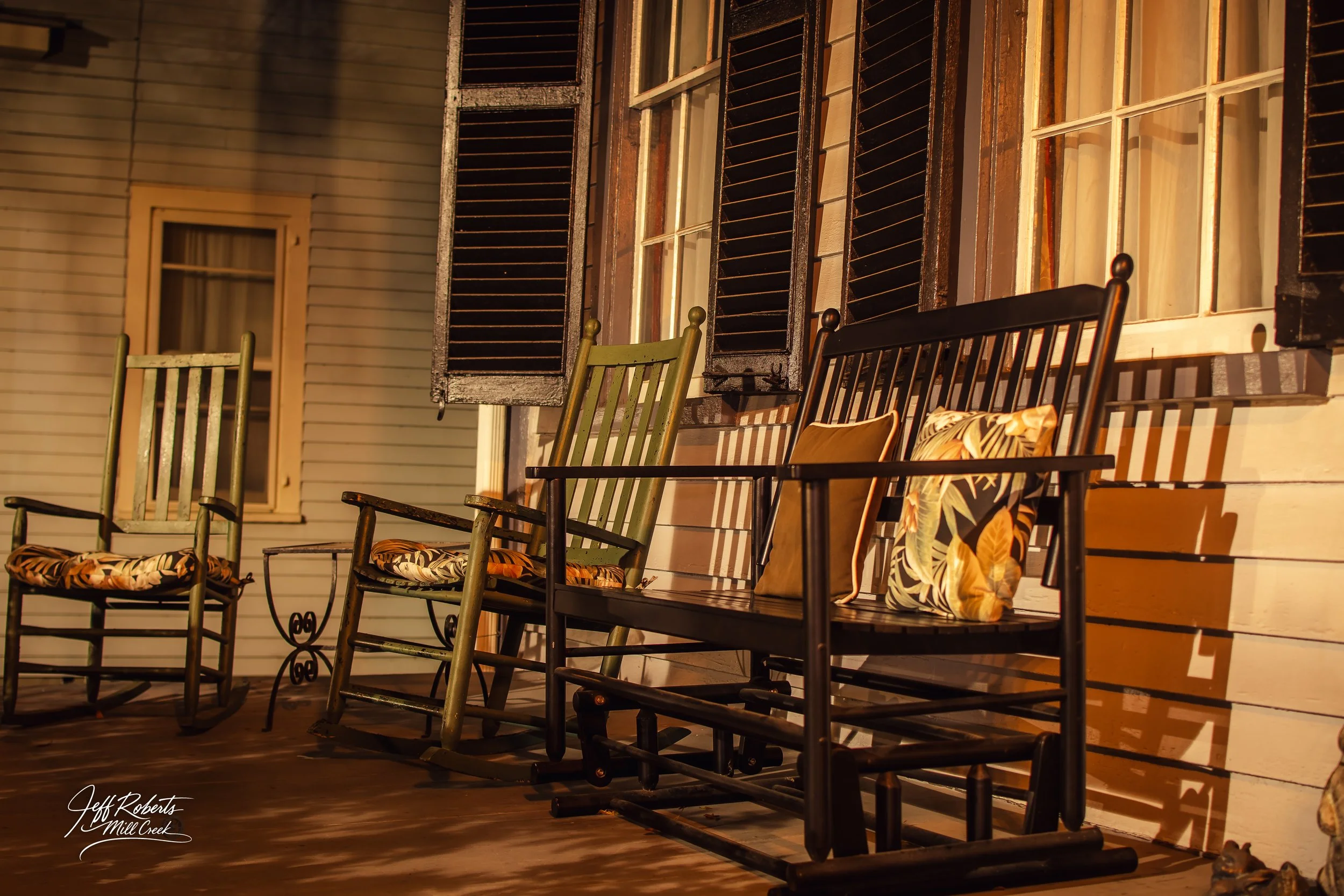 Porch with four rocking chairs with floral and solid cushions, in front of a house with white wooden siding and open dark brown window shutters, illuminated by warm yellow lighting at night.