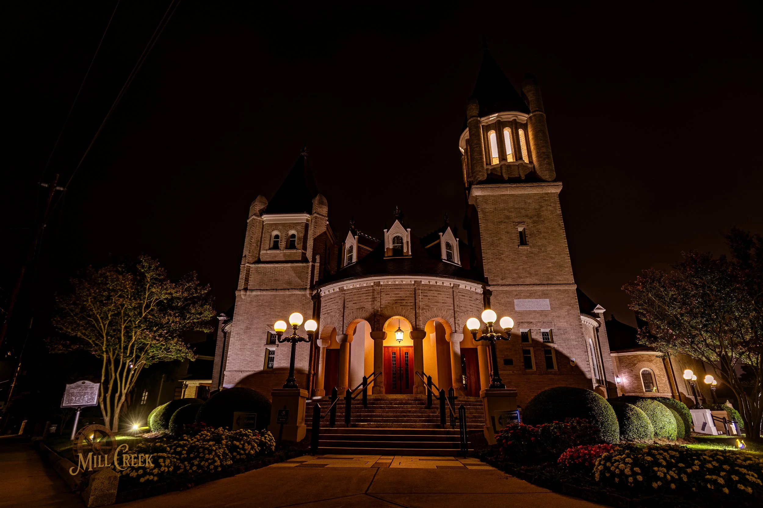 A large, historic church illuminated at night with a front entrance, stairs, and surrounding landscaped grounds with bushes and trees.