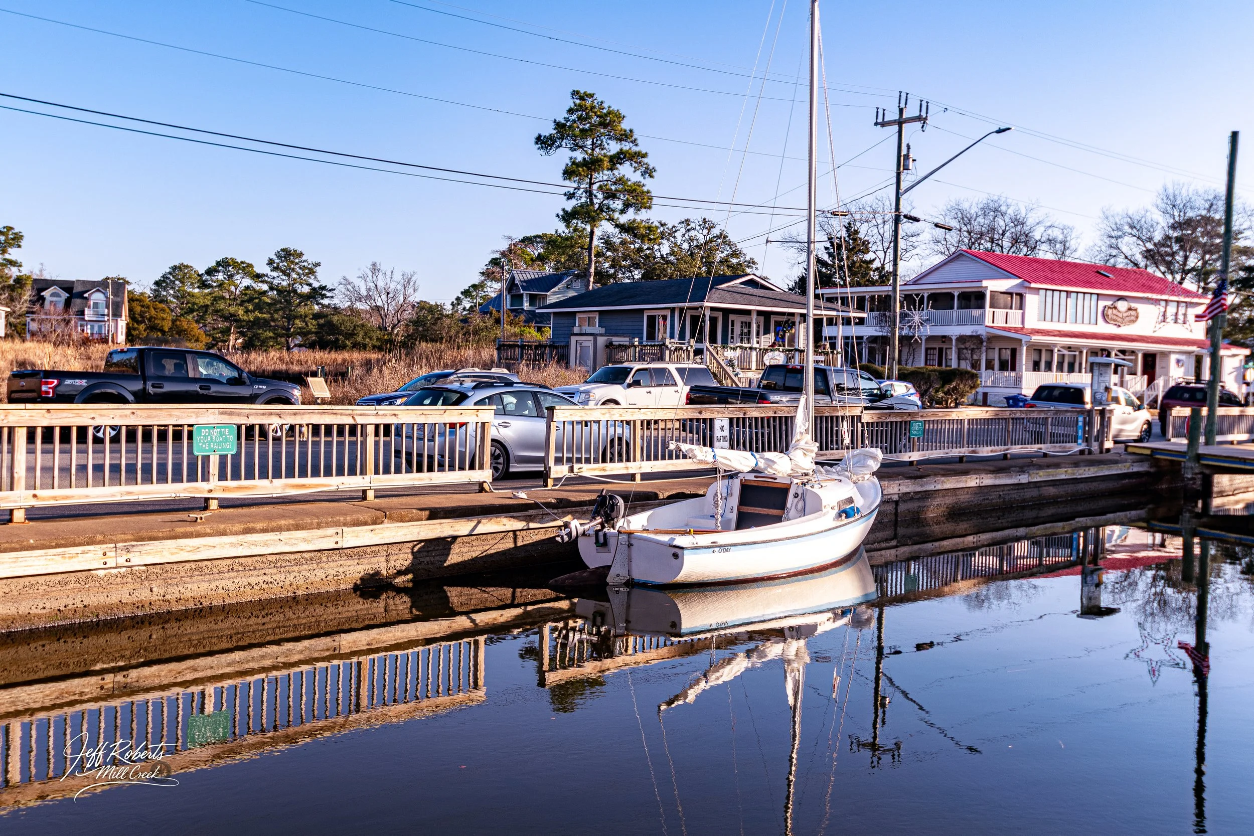 A small white sailboat docked in a marina with calm water, surrounded by houses and parked cars on a clear day.