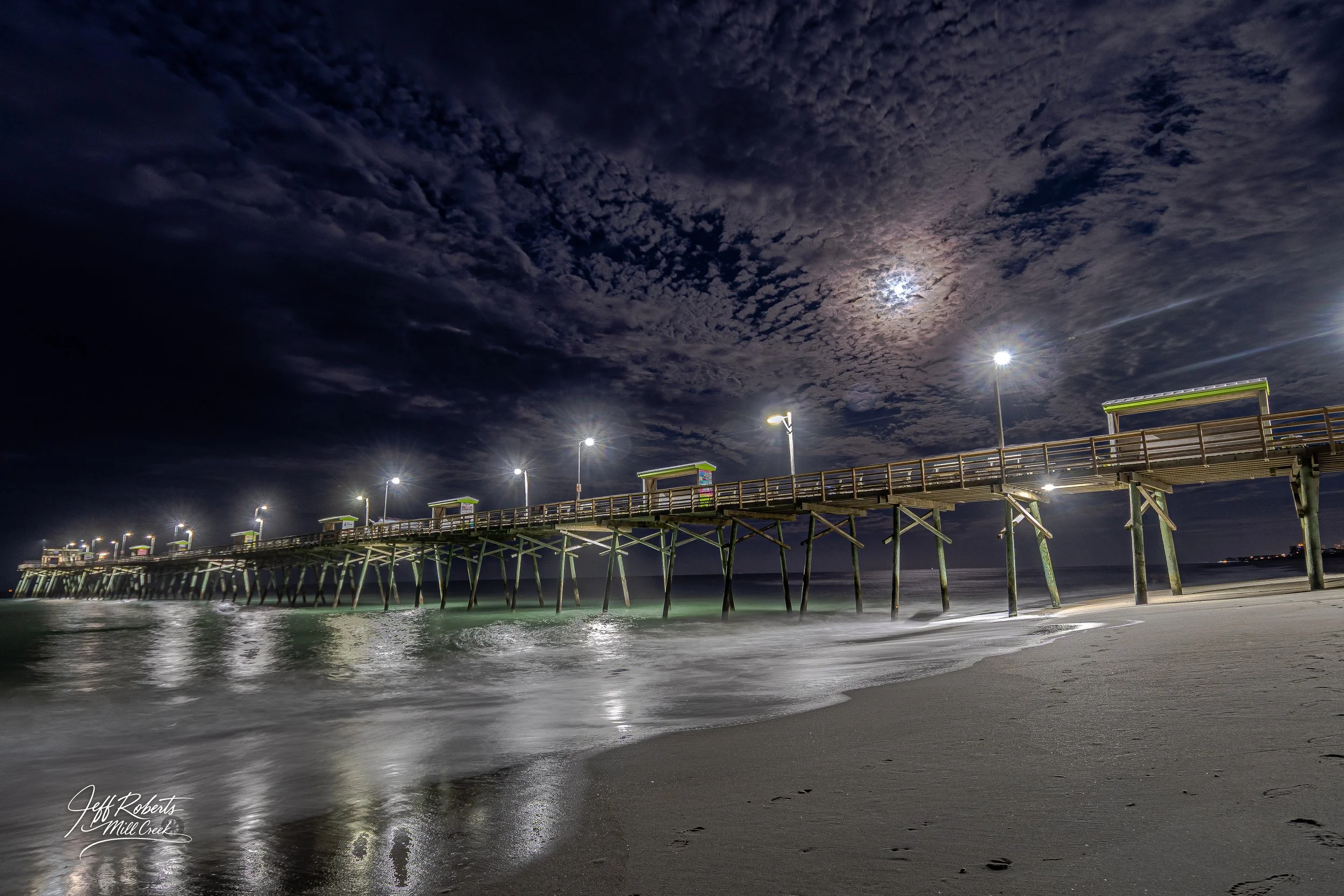 Nighttime view of a pier with wooden supports extending into the ocean, illuminated by streetlights, with a cloudy sky and moon in the background.