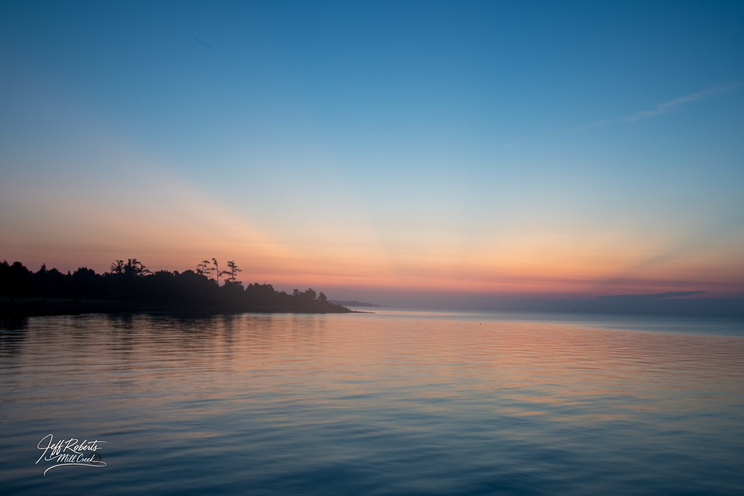 Sunset over a calm river with silhouetted trees on the shoreline and a colorful sky with shades of pink, orange, and blue.