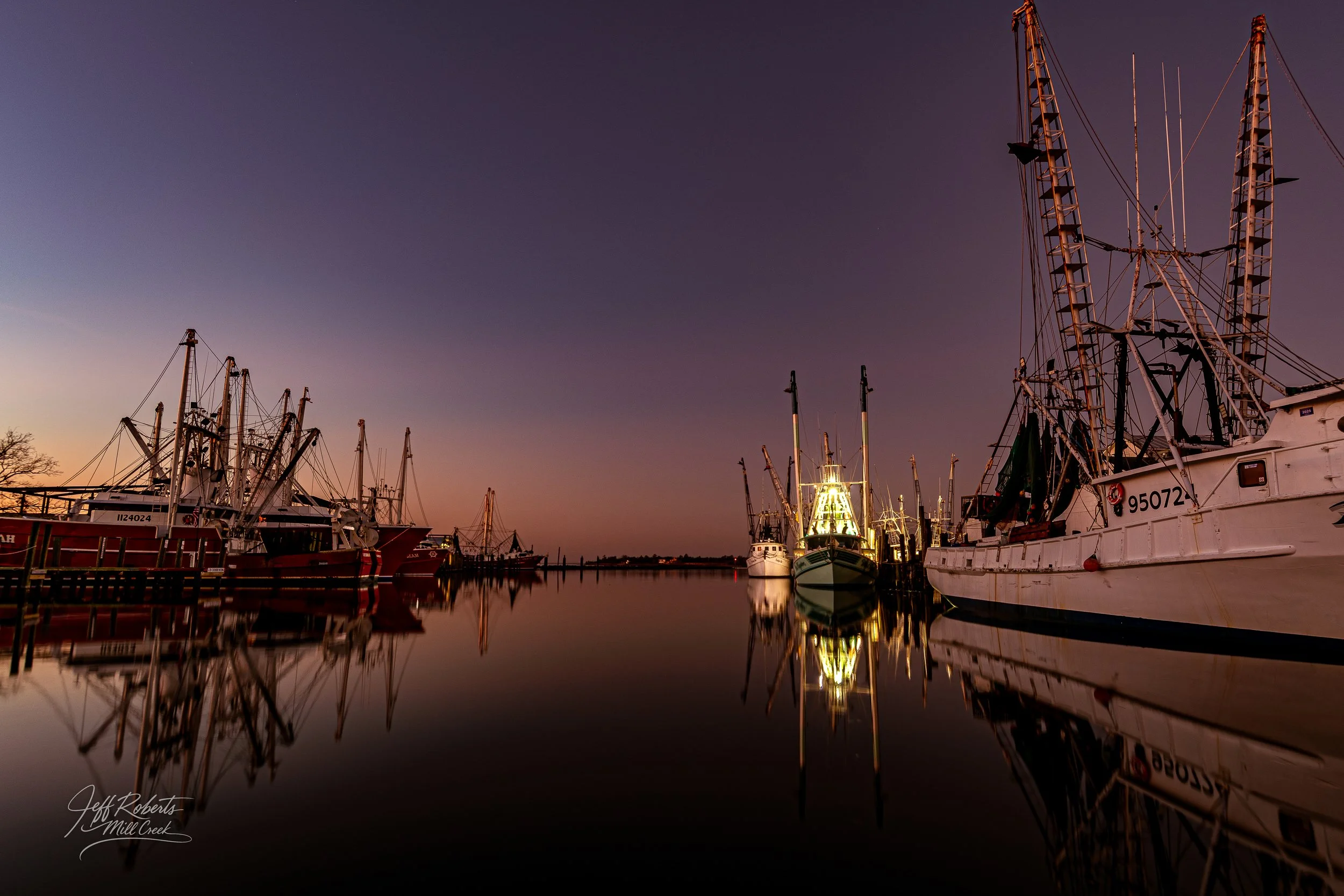 Boats docked at a harbor during sunset with calm water reflecting the vessels and a colorful sky.