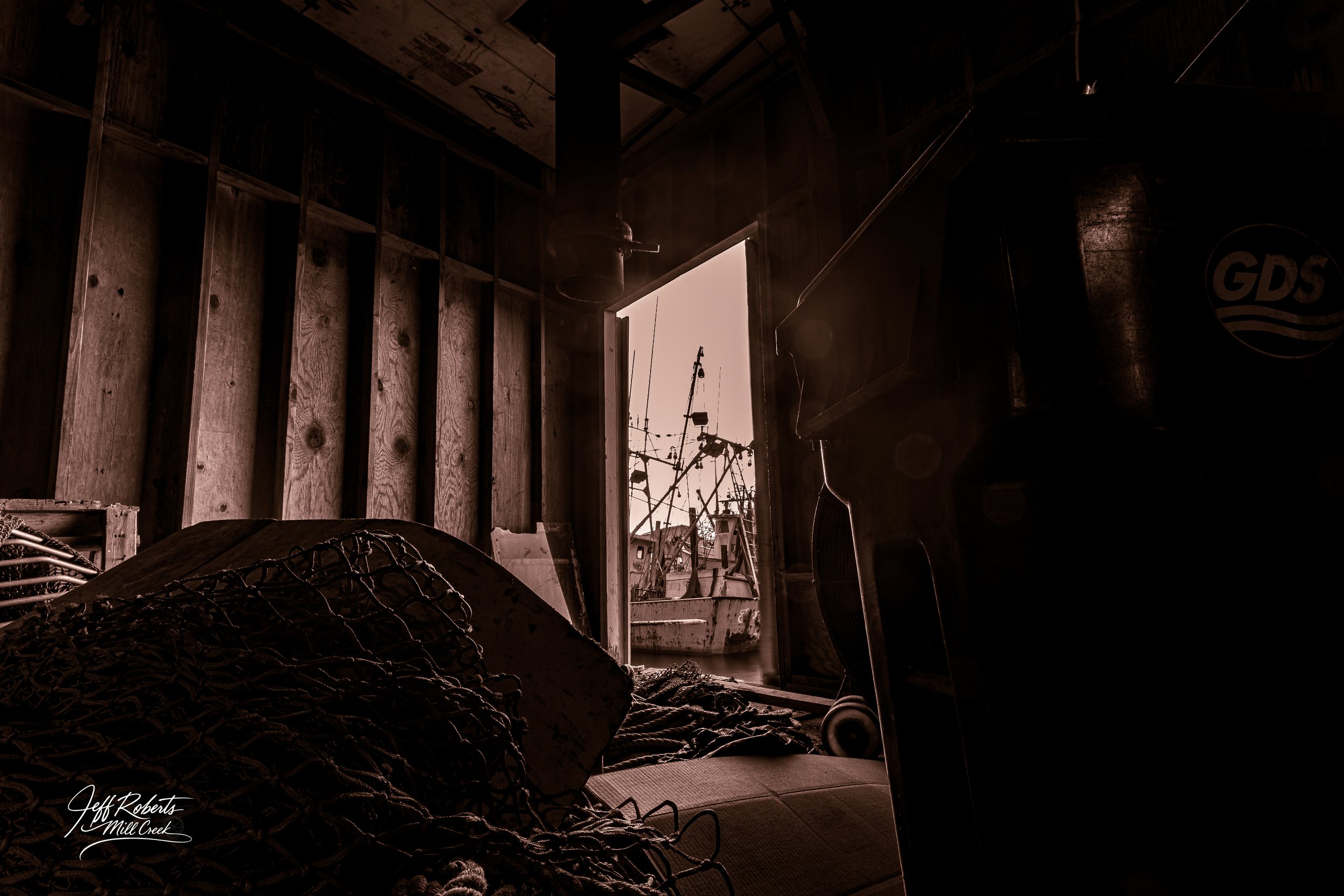 Interior of a boat shed with fishing nets and equipment, open door showing moored fishing boats outside