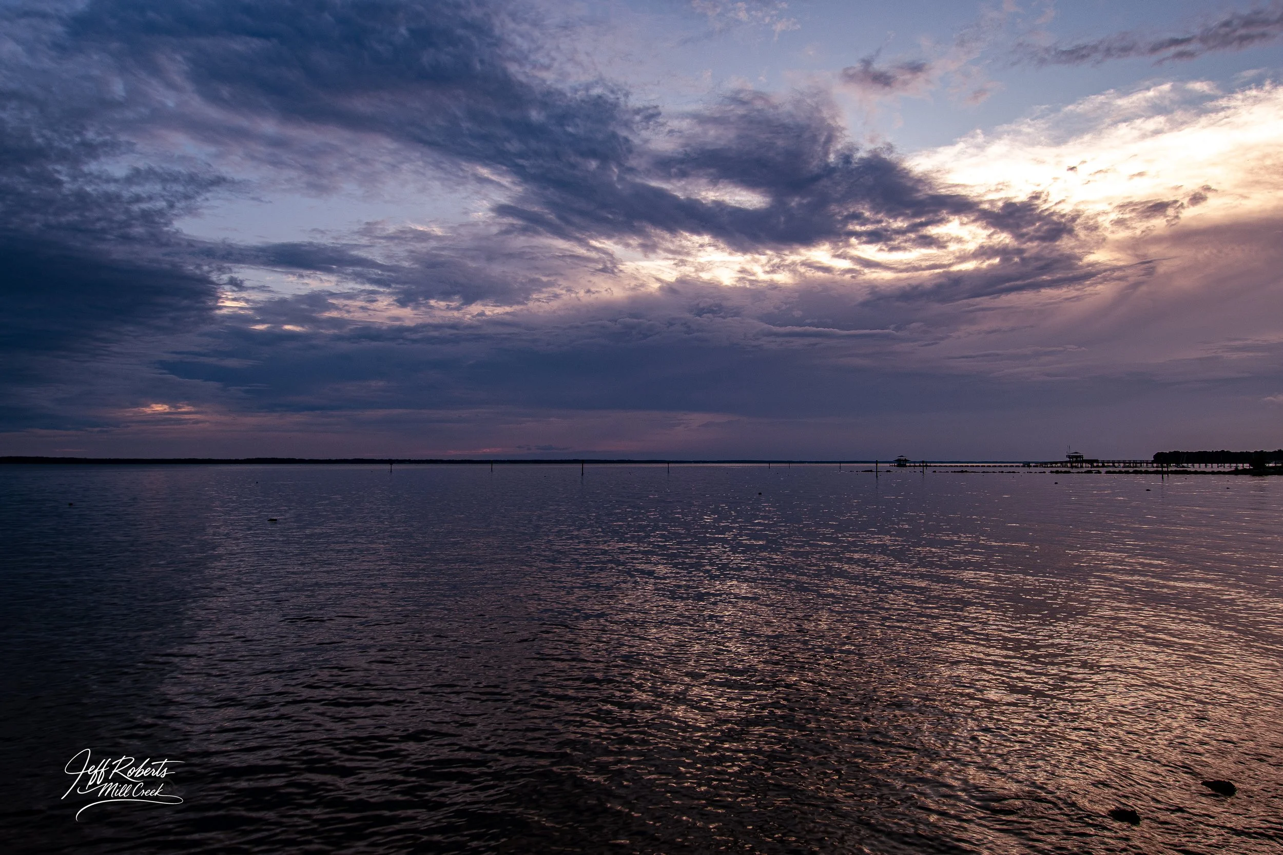A scenic view of a large body of water at sunset, with a partly cloudy sky and some structures or docks in the distance.