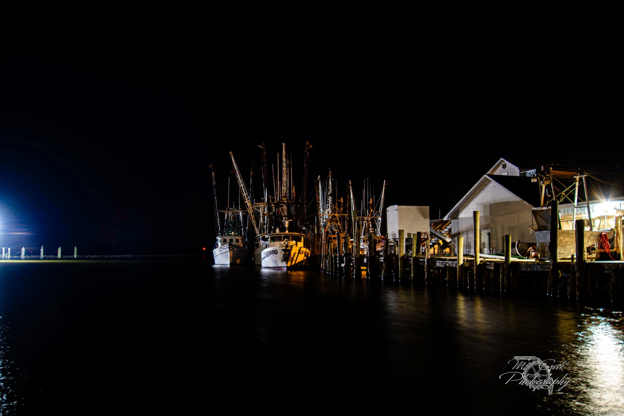 "Night Harbor Glow"
Fishing boats rest quietly at the dock under a blanket of night, their masts and rigging silhouetted against the dark sky while bright dock lights spill reflections across the still water.