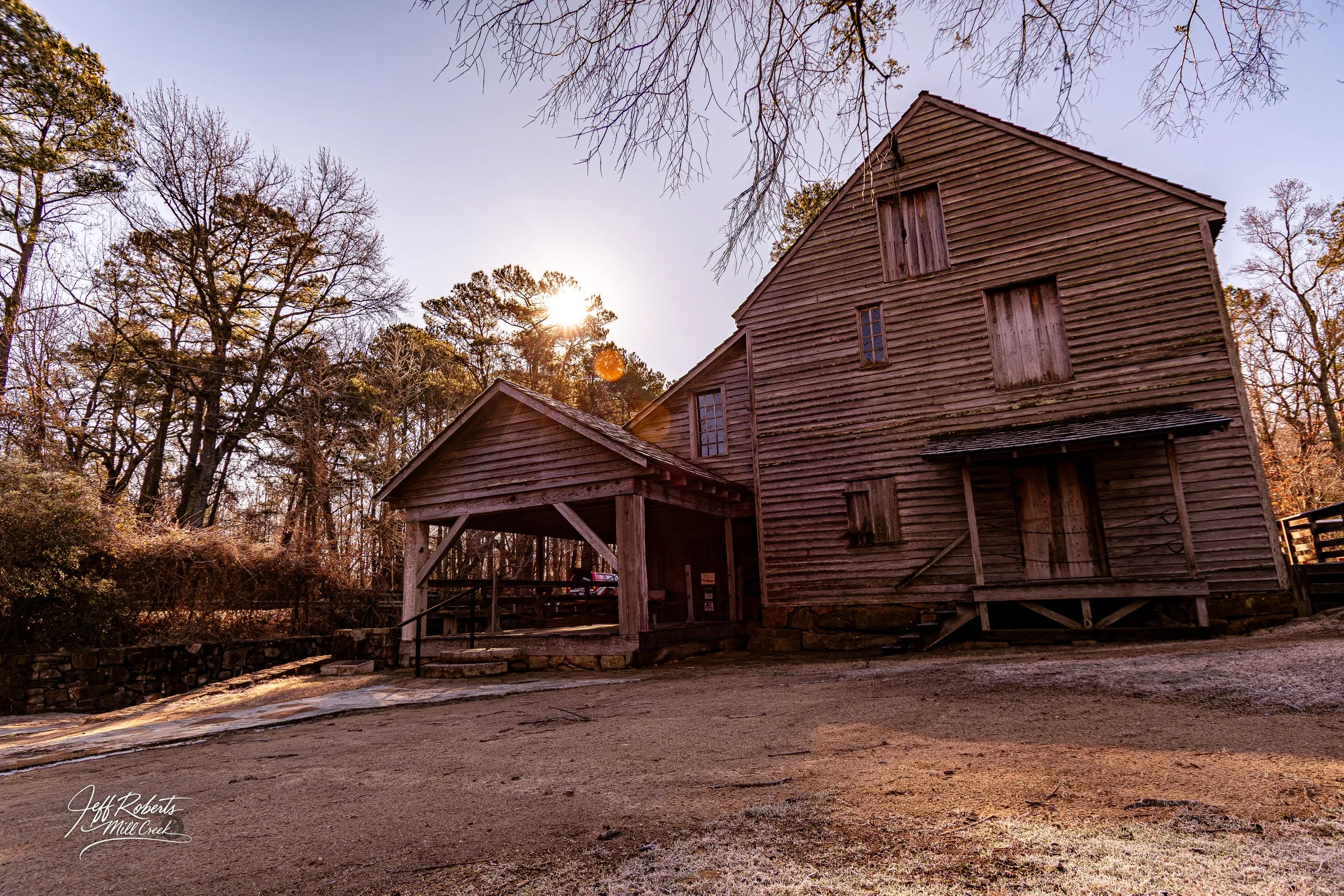 An old wooden barn with boarded-up windows and a small covered porch, surrounded by leafless trees and a dirt ground, with the sun setting behind the trees.