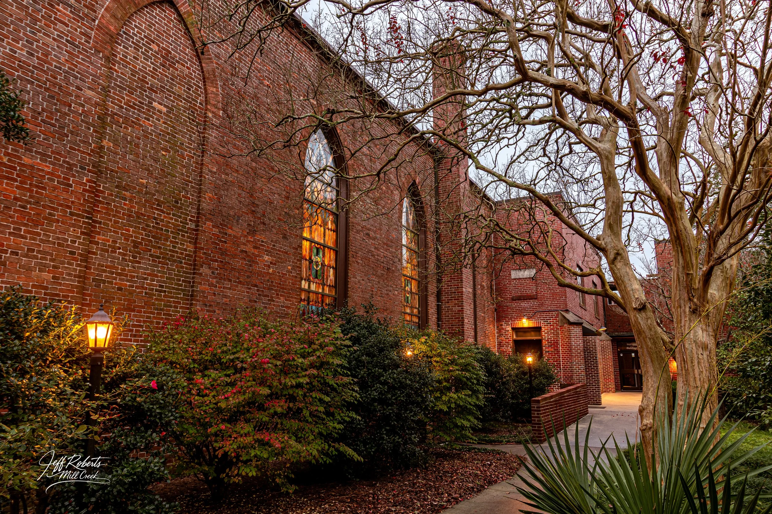 A brick church with stained glass windows and leafless trees outside, lit by street lamps during dusk.