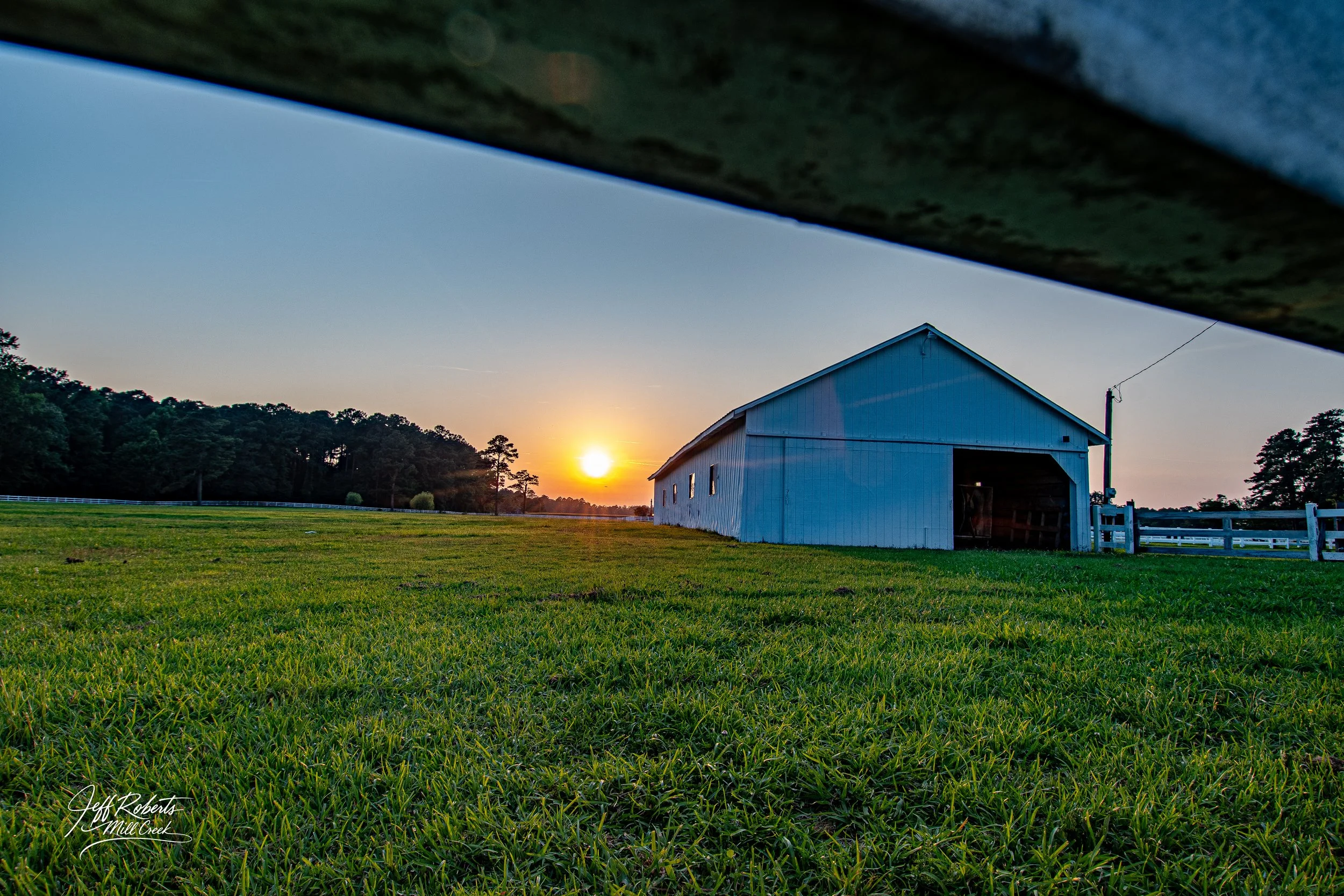A rural scene at sunset featuring a white barn on lush green grass with a backdrop of trees and a clear sky. The photo is taken from beneath a shaded overhang.
