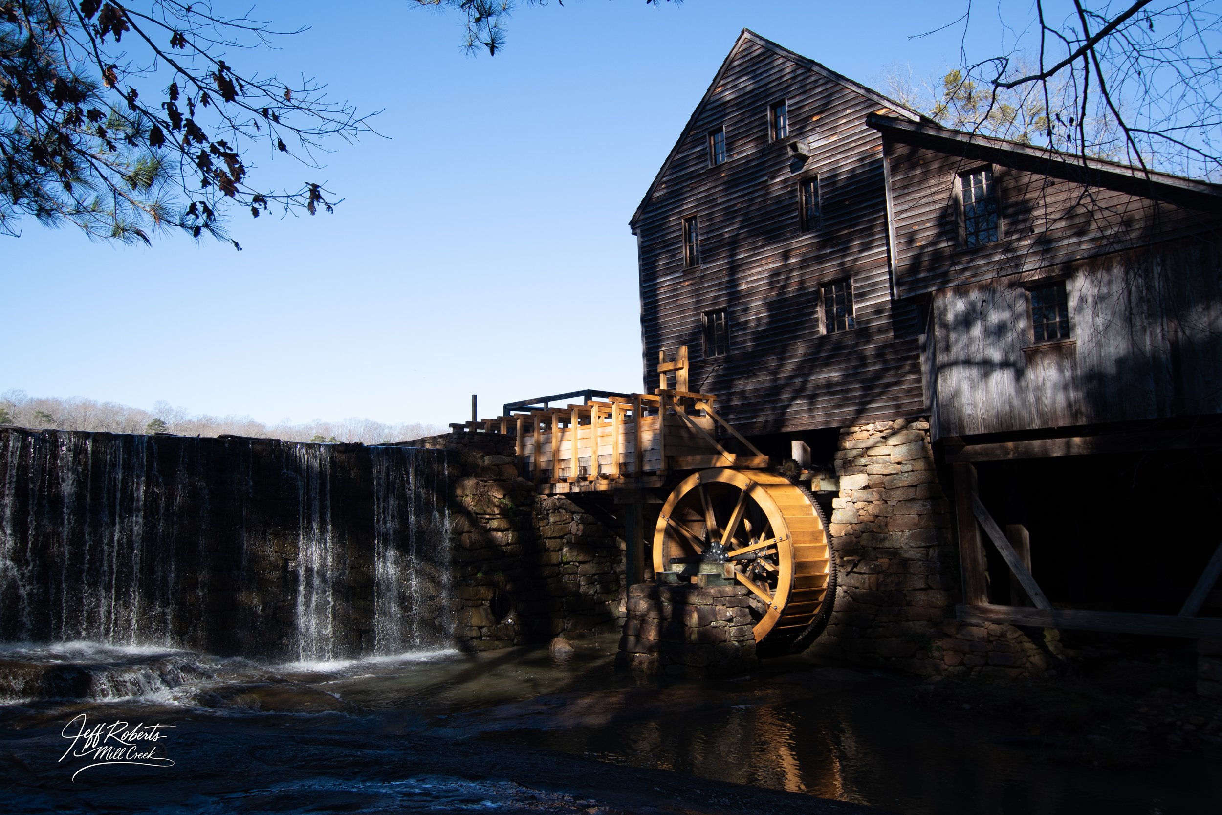 An old wooden watermill next to a small waterfall with flowing water, under a clear blue sky, with shadows of trees on the building.