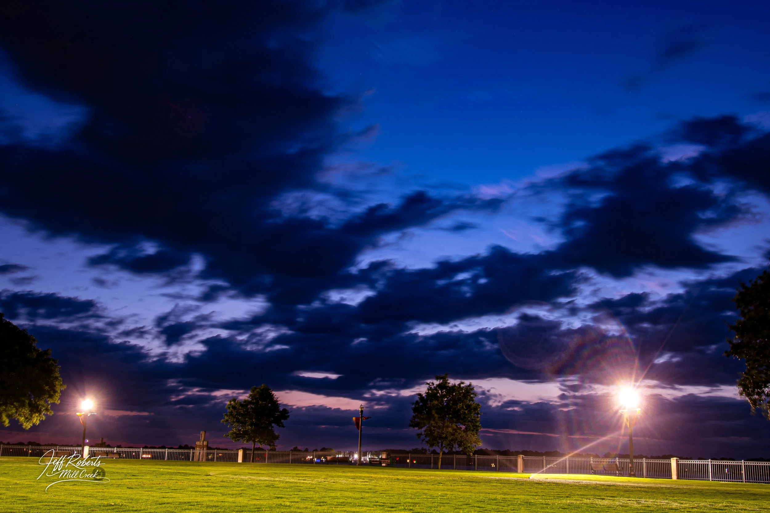 Nighttime park with green grass, trees, and bright streetlights under a mostly cloudy dark blue sky.
