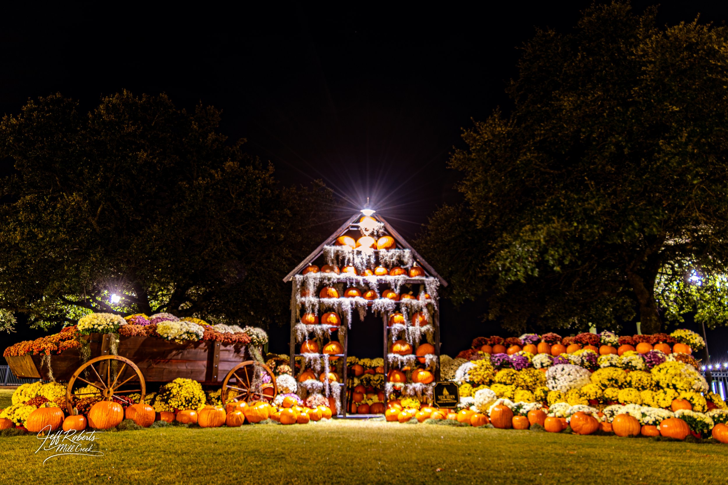 Night scene of a pumpkin display with a wooden cart filled with flowers on the left, a wooden arch with stacked pumpkins in the center, and more pumpkins and flowers on the right, all illuminated by artificial lights under a dark sky.