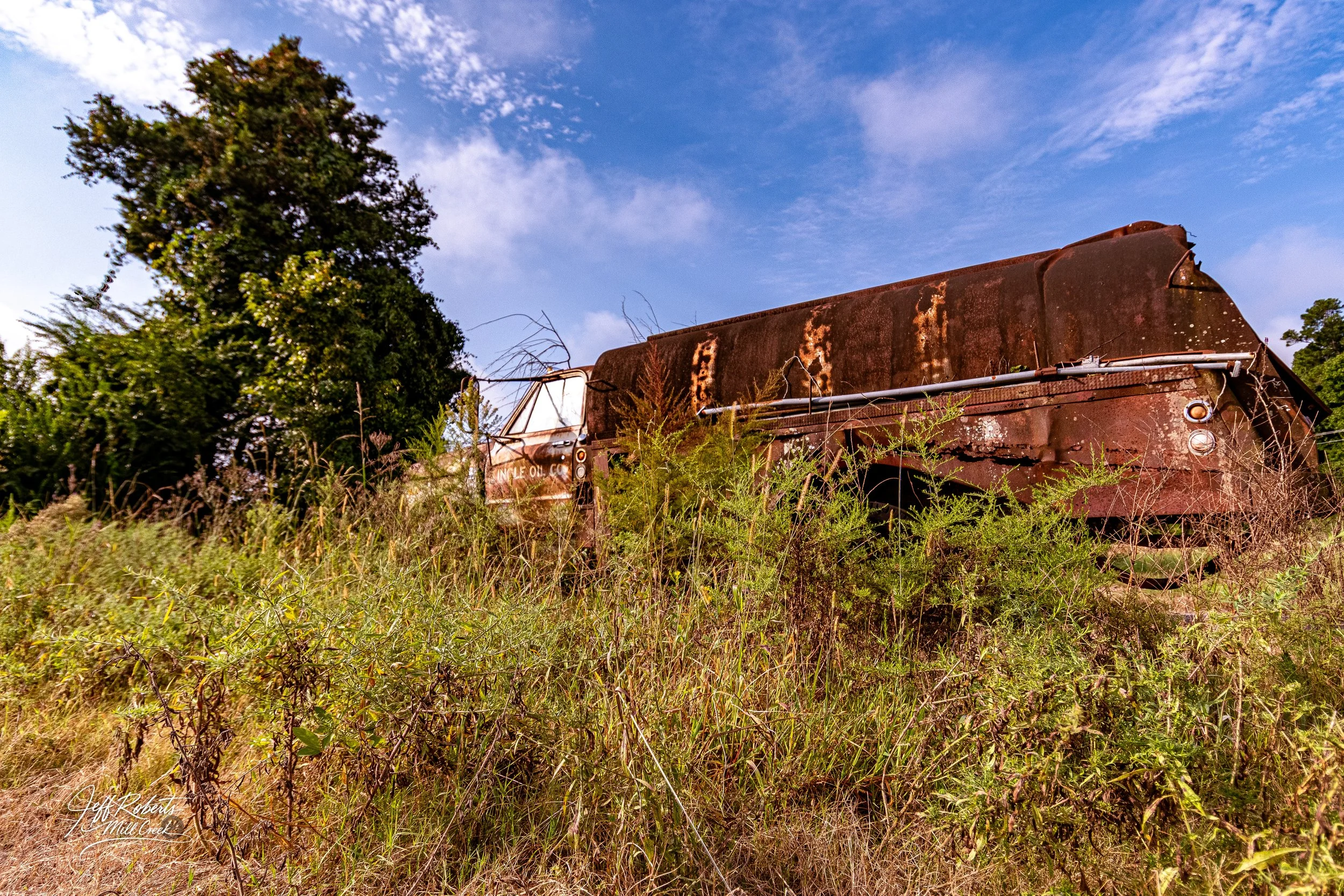 Old rusted oil truck partially obscured by tall grass and plants, against a background of blue sky and scattered clouds.