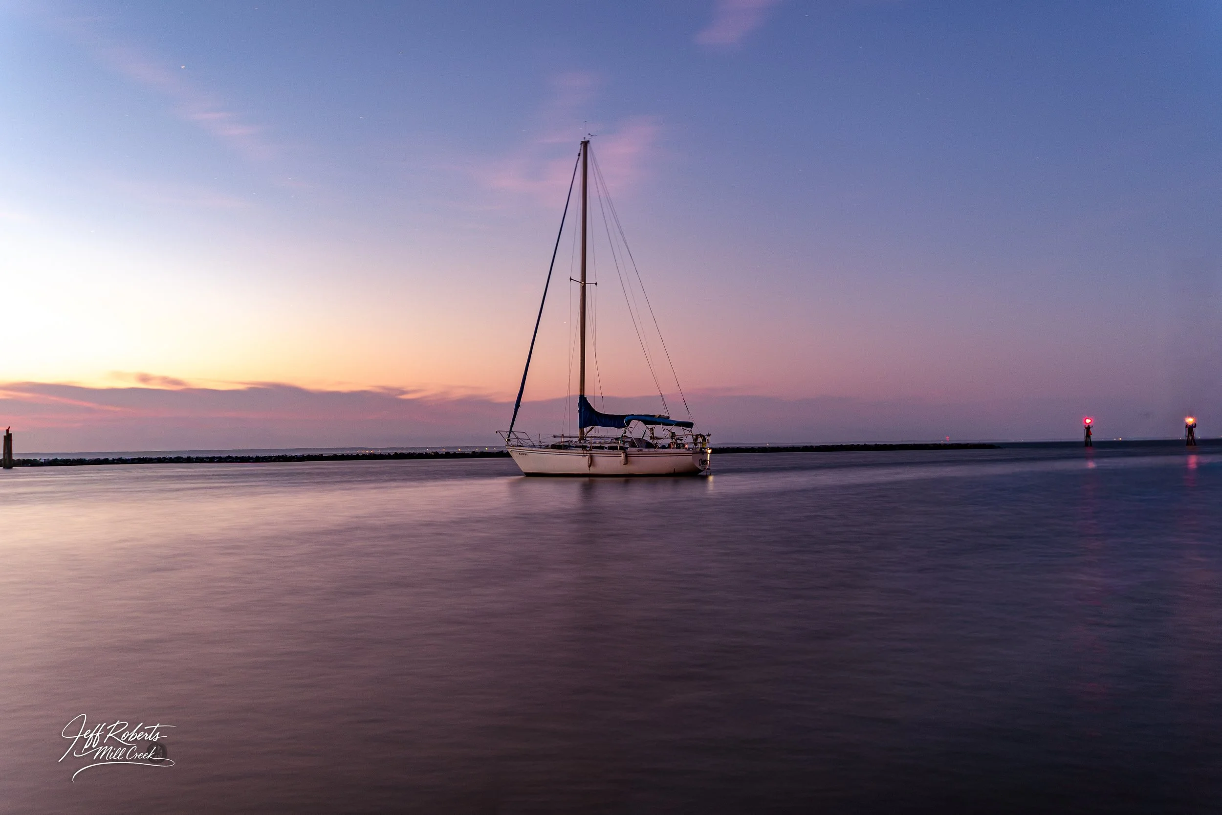 A sailboat anchored in calm waters during twilight, with a colorful sky and distant navigation lights.