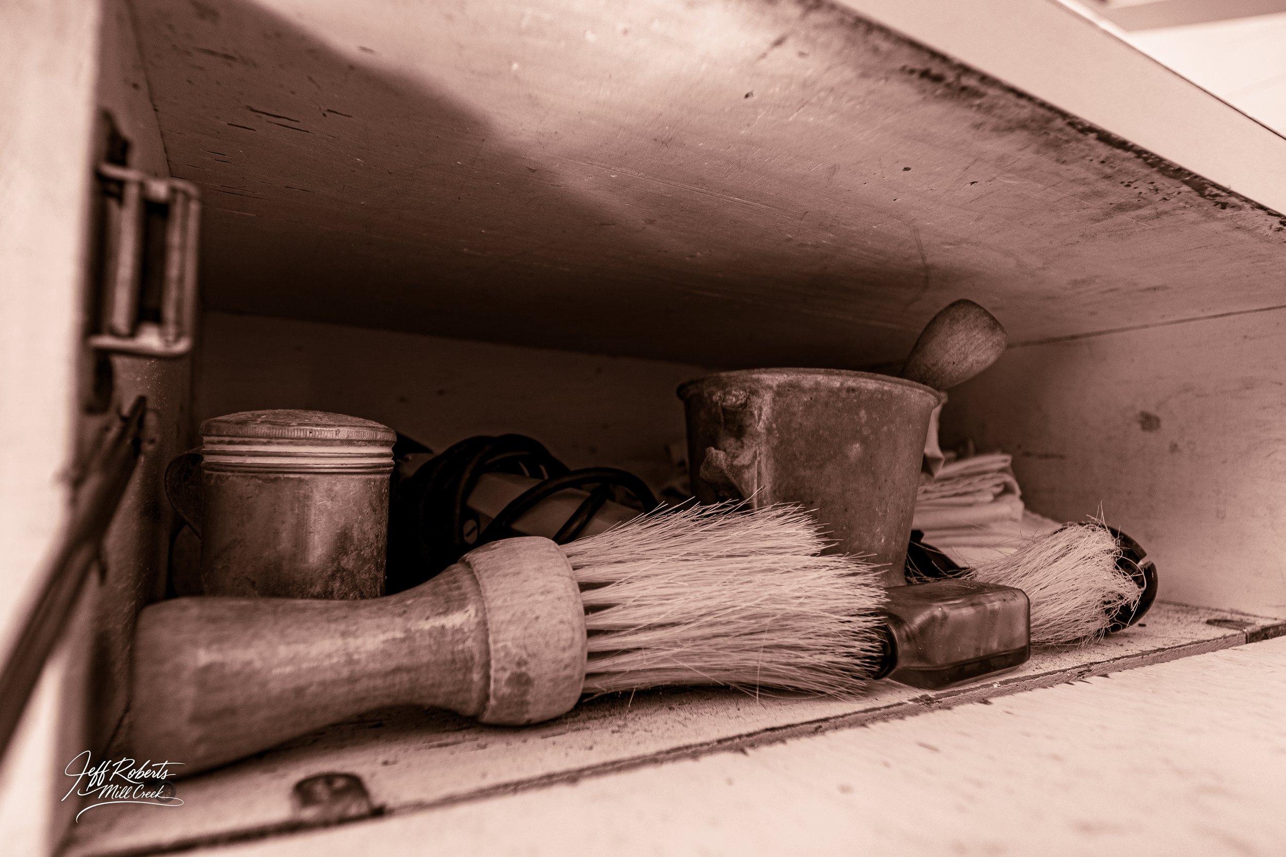 A dusty kitchen drawer with a wire whisk, a candle tin, a small metal pot, a pipe, a small container, and some papers.