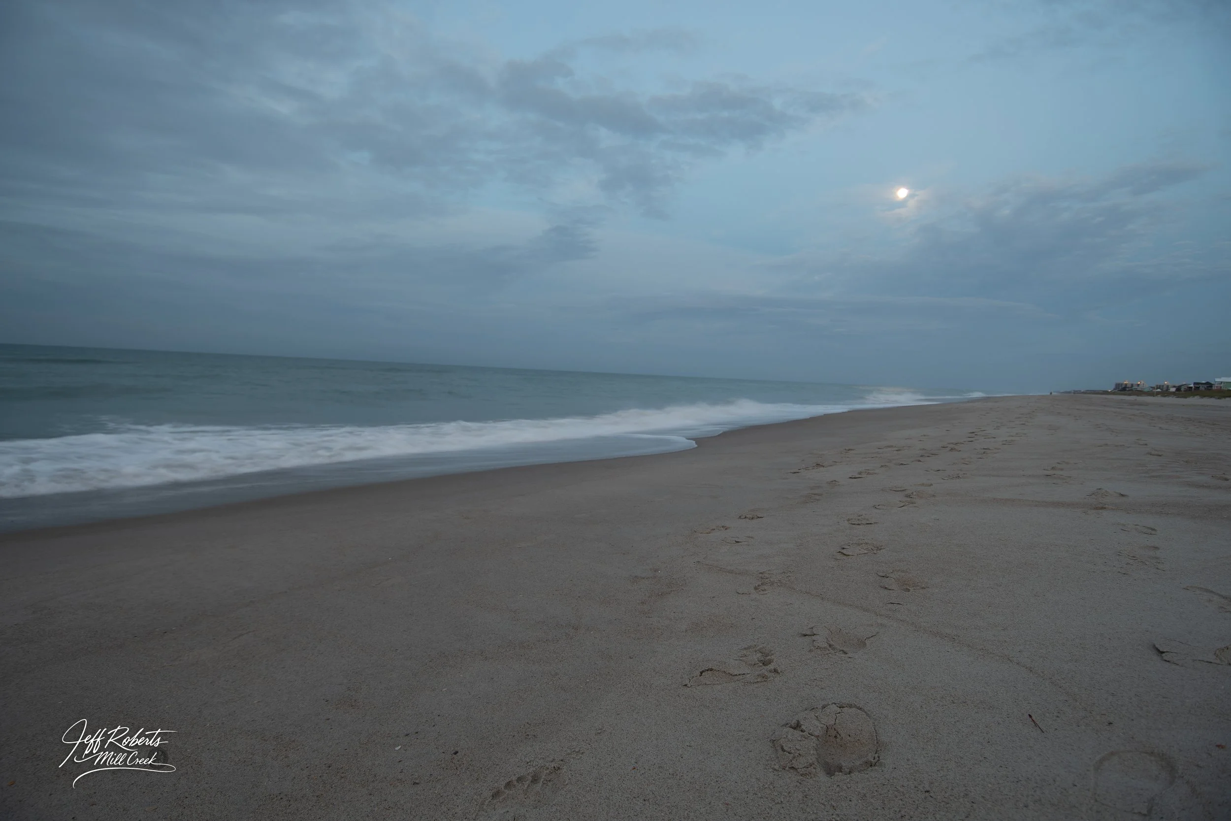 Empty beach with footprints in the sand during twilight, waves gently crashing onto the shore, cloudy sky with the moon partially visible.