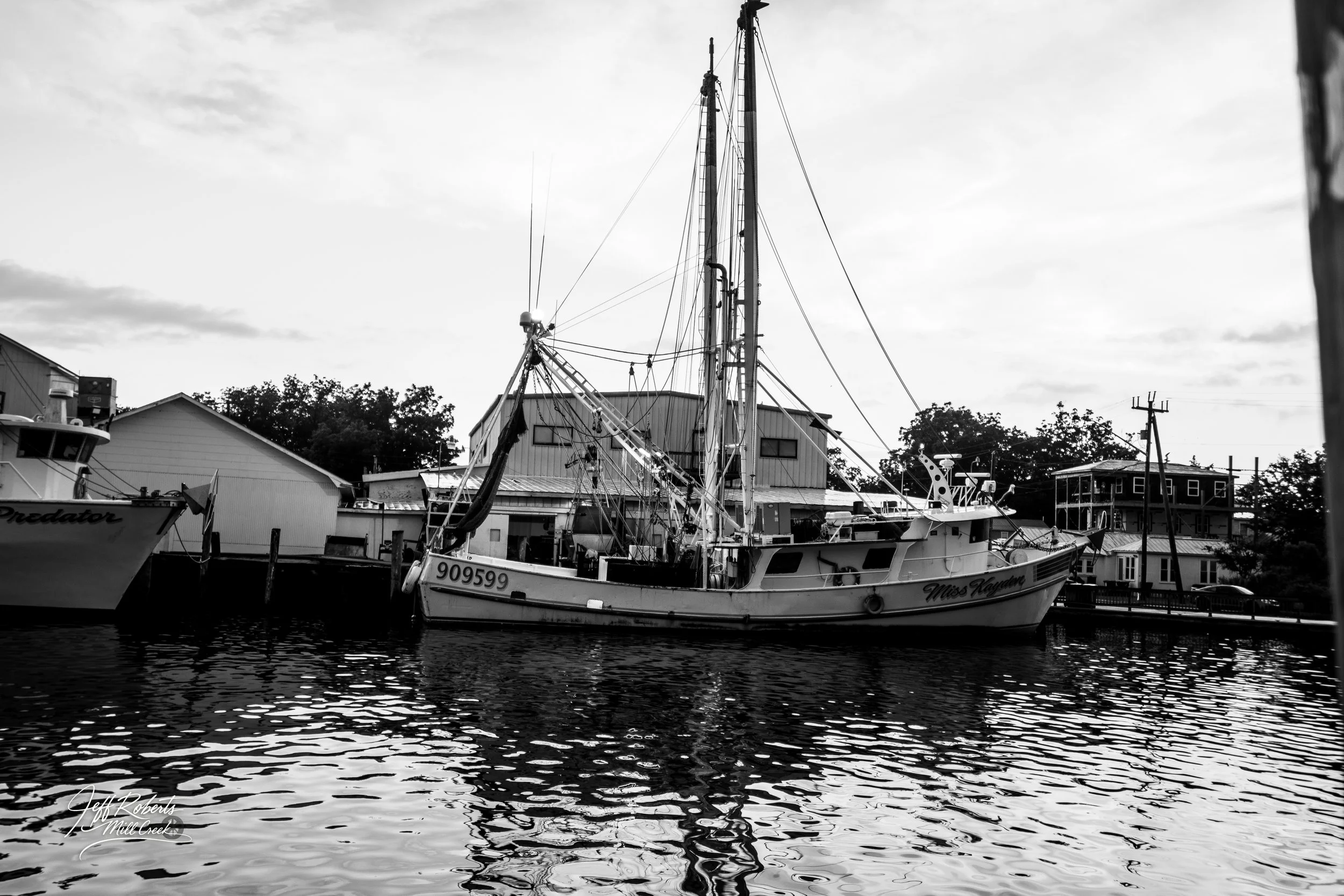 Black and white photo of boats docked at a marina with buildings in the background.