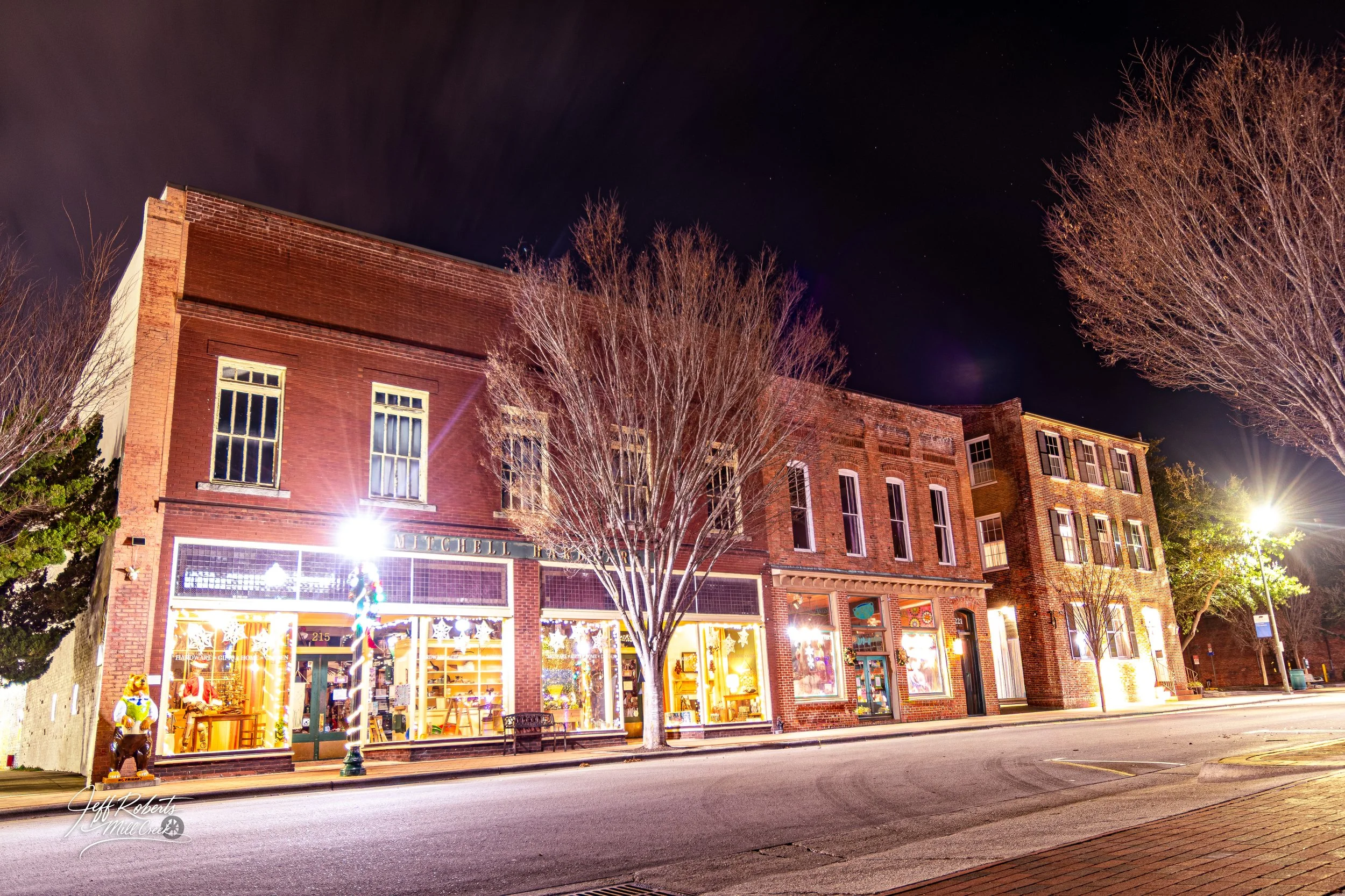 Nighttime street view of a brick building with store windows, trees, and streetlights illuminating the scene.