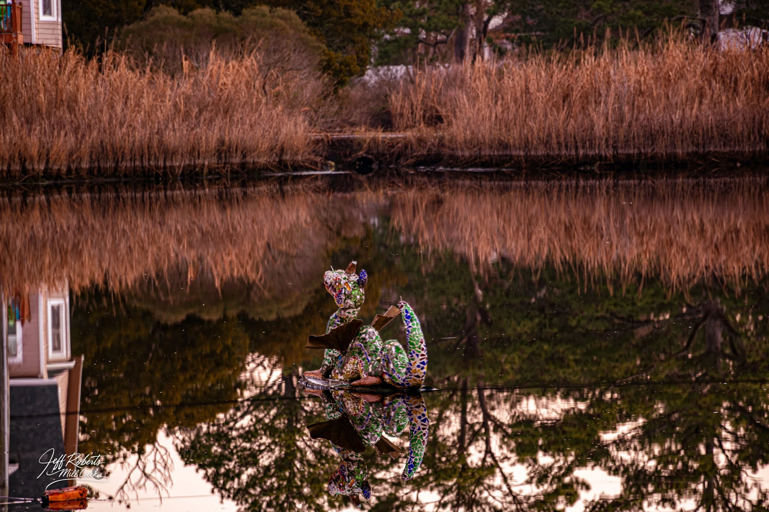 Colorful mosaic sculpture of a sitting animal, possibly a fox, reflected in a calm body of water surrounded by brown reeds and trees.