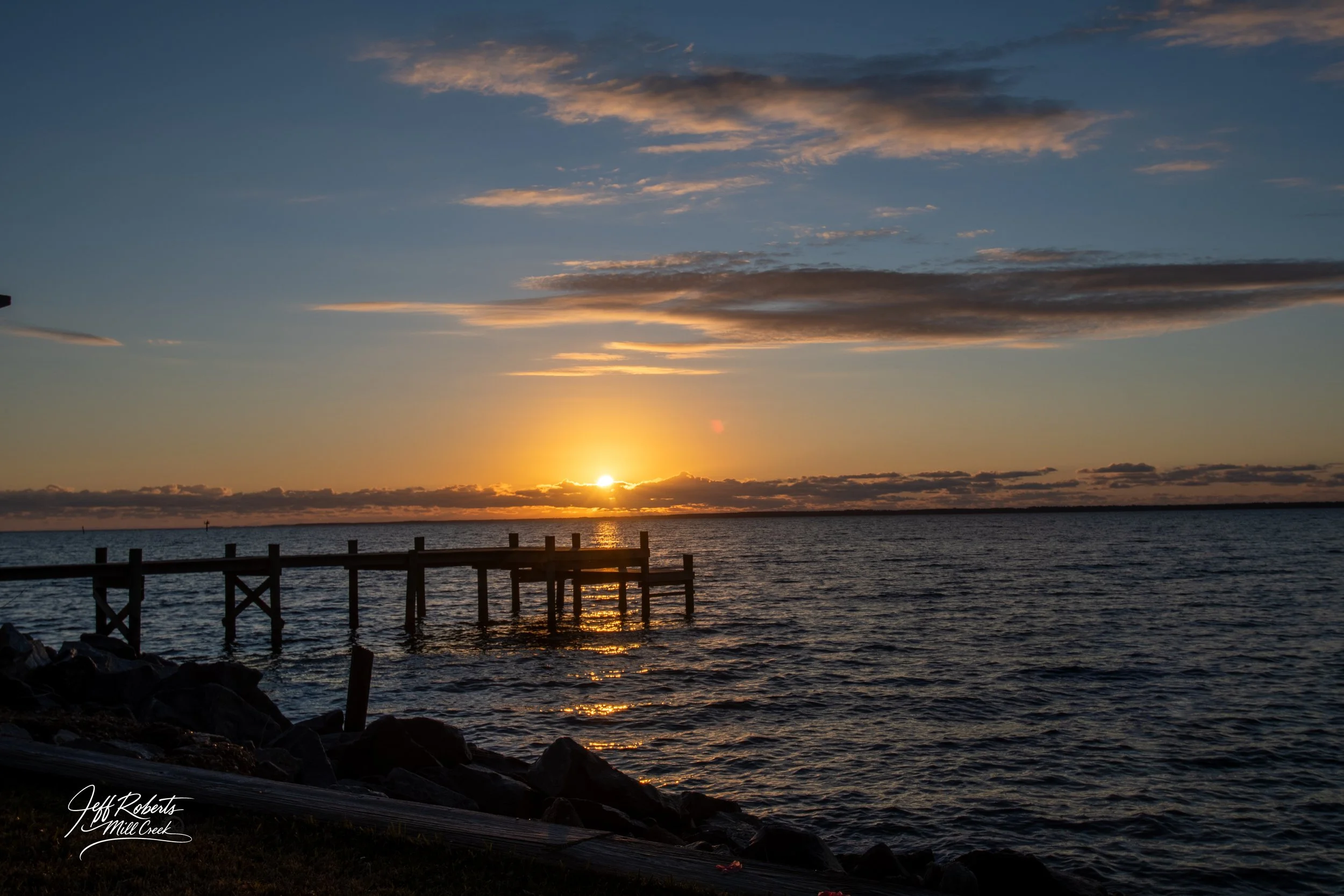Sunset over a body of water with a wooden pier extending into the water and a rocky shoreline in the foreground.